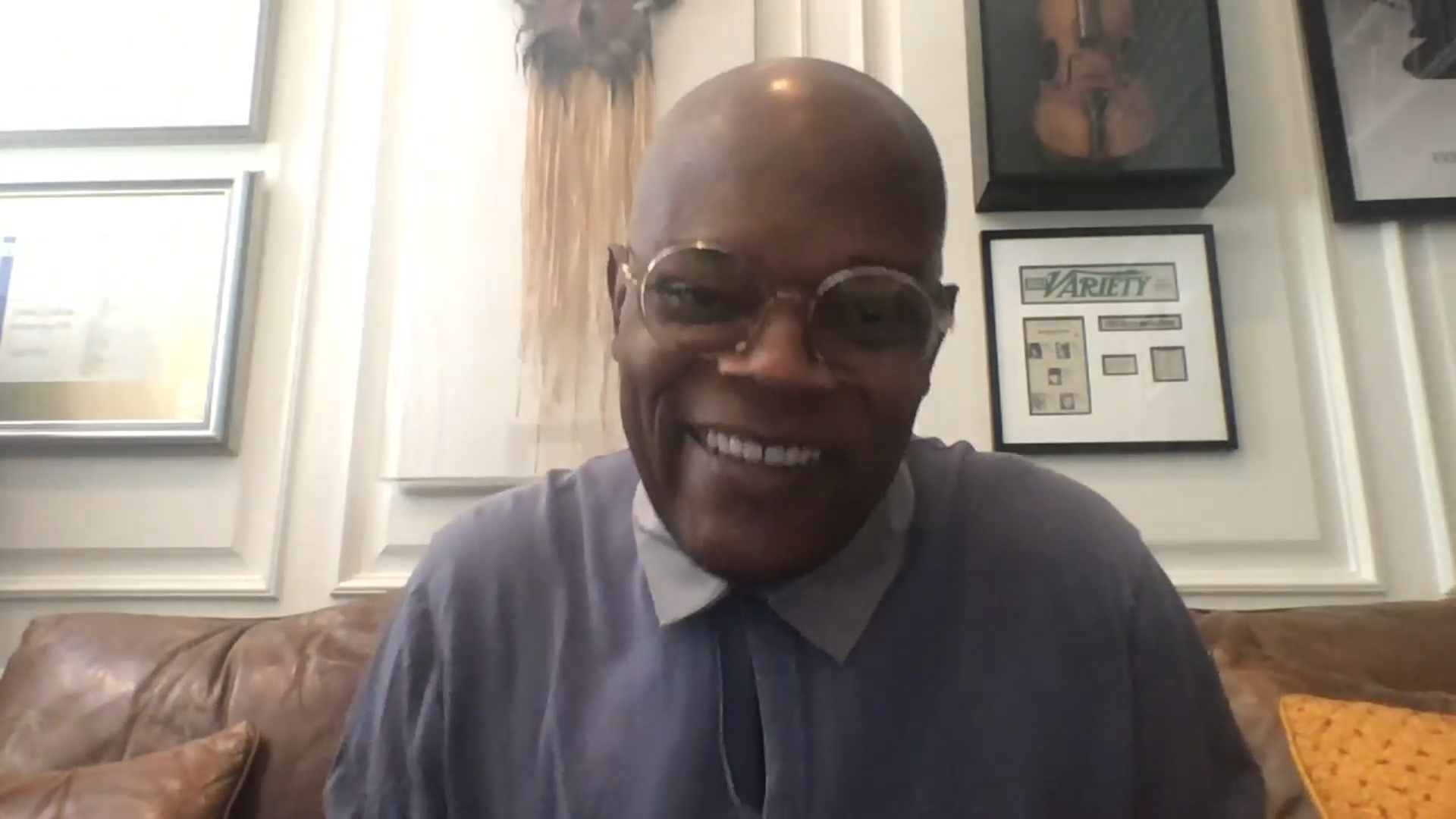 A smiling man with glasses, a bald head, wearing a light-colored shirt and a tie, seated on a brown leather couch in a room with framed art and newspaper prints on the wall.