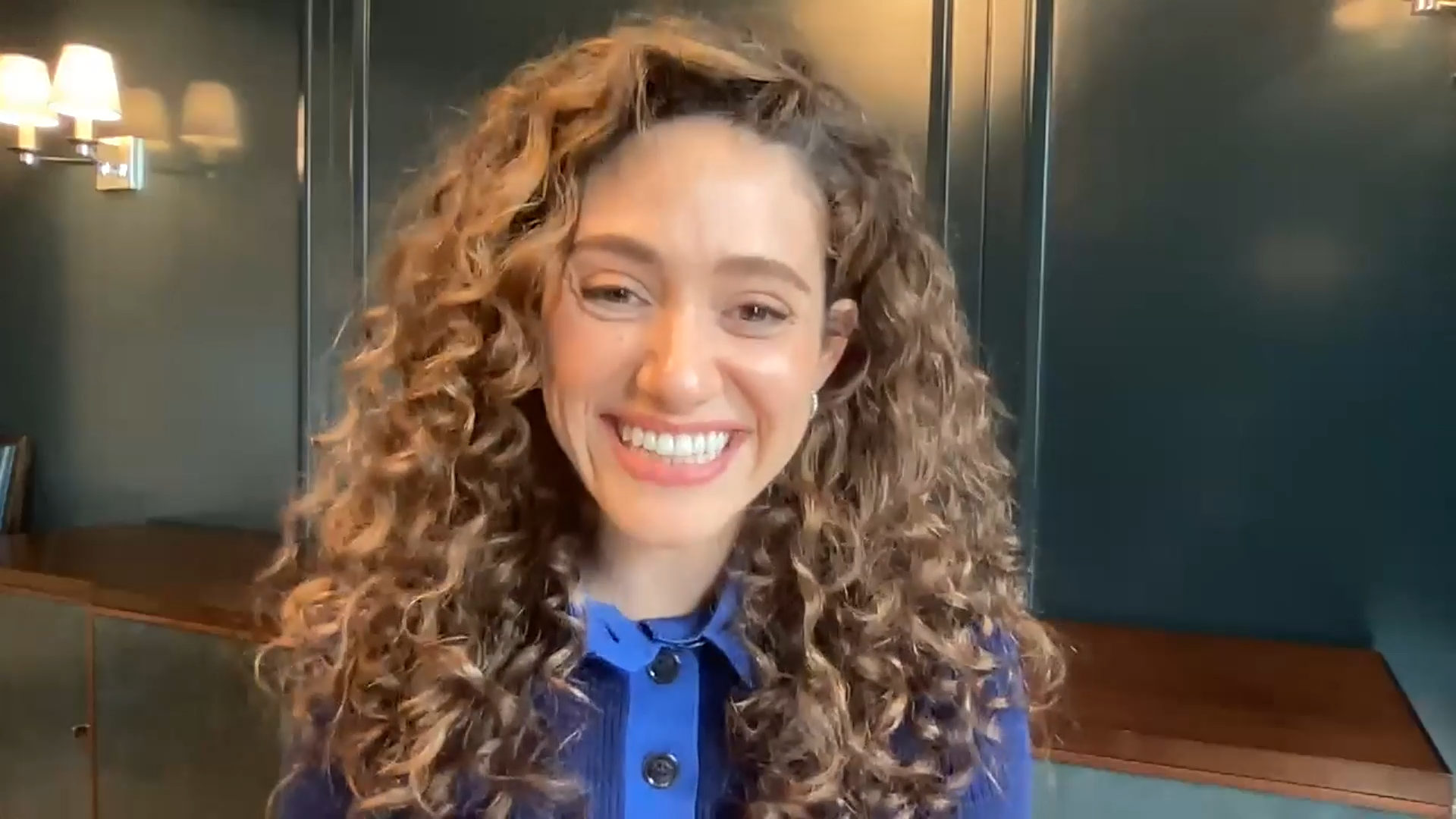 Close-up of a woman with long, curly brown hair, wearing a blue top, smiling in an indoor setting with dark green walls and warm lighting.