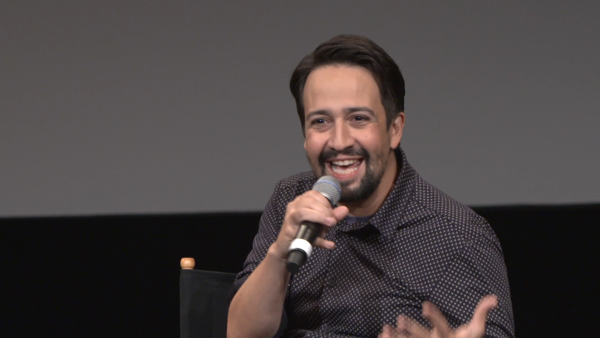 A man with dark hair and a beard, smiling, holding a microphone, speaking at an event.