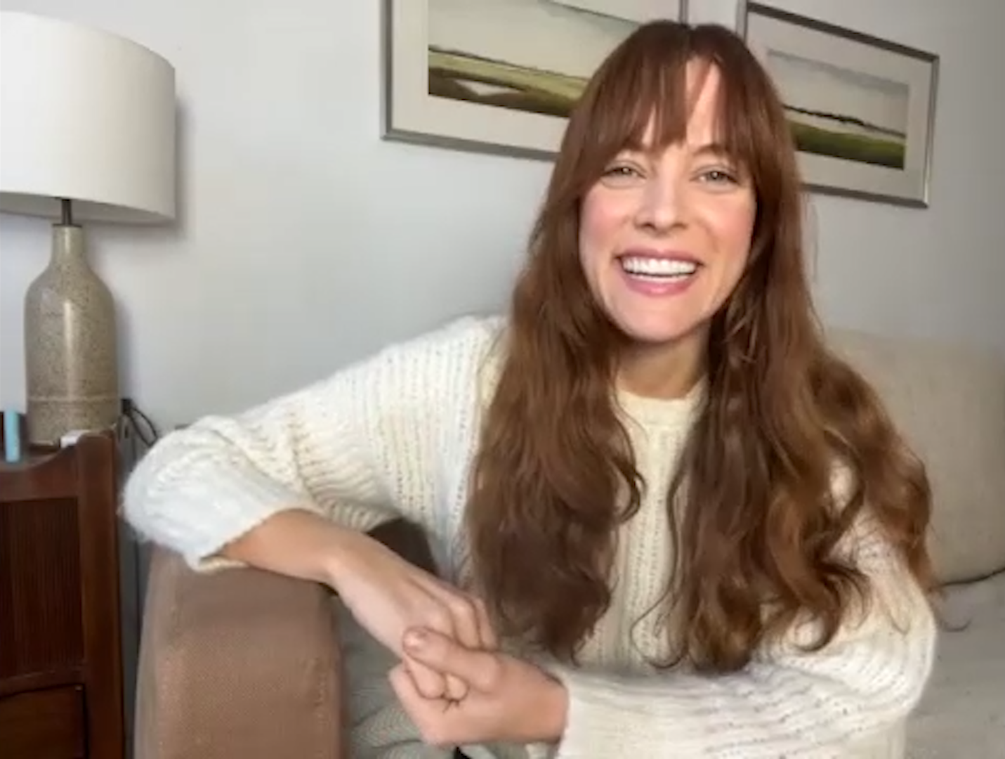 A woman with long, wavy red hair smiling while sitting on a beige couch in a living room. She is wearing a cream-colored knit sweater, and her left arm rests on the armrest of the couch. Behind her is a wall with framed landscape artwork and a side table with a ceramic lamp.