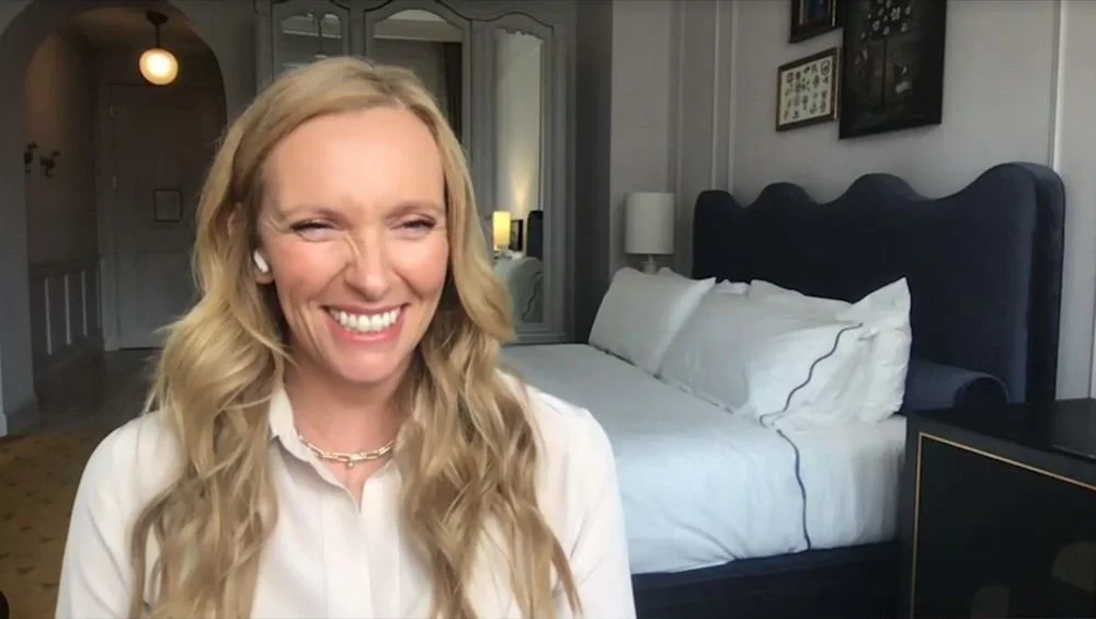 A woman with long blonde hair smiling, sitting in a hotel room with a bed and framed pictures on the wall behind her.
