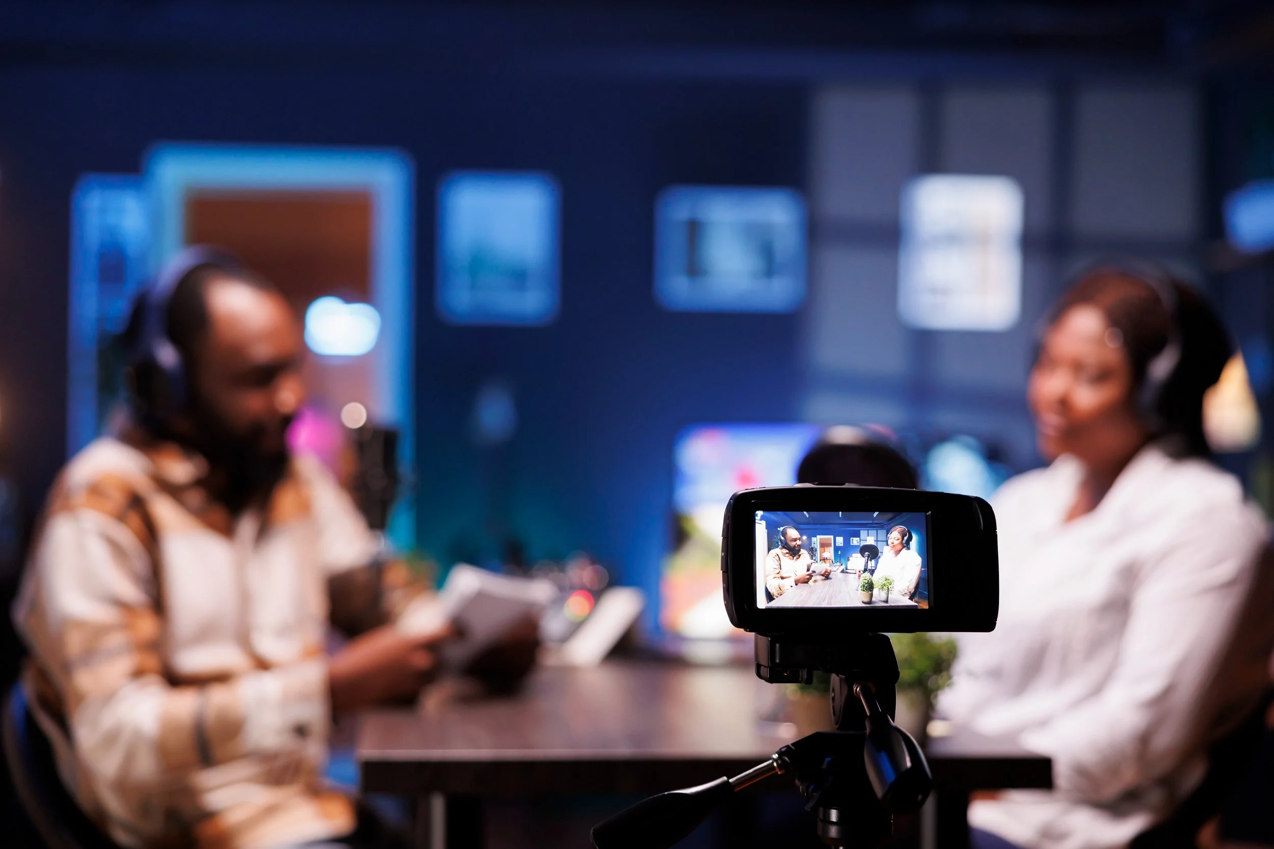Two women with headphones, sitting at a table during a recording session or interview, with a camera in the foreground capturing the scene.