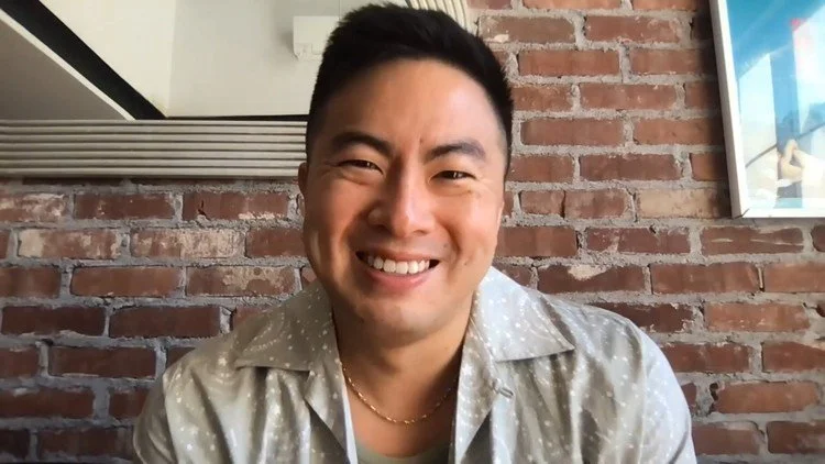 A smiling man with short black hair and a light patterned shirt, sitting in front of a brick wall in a casual setting.