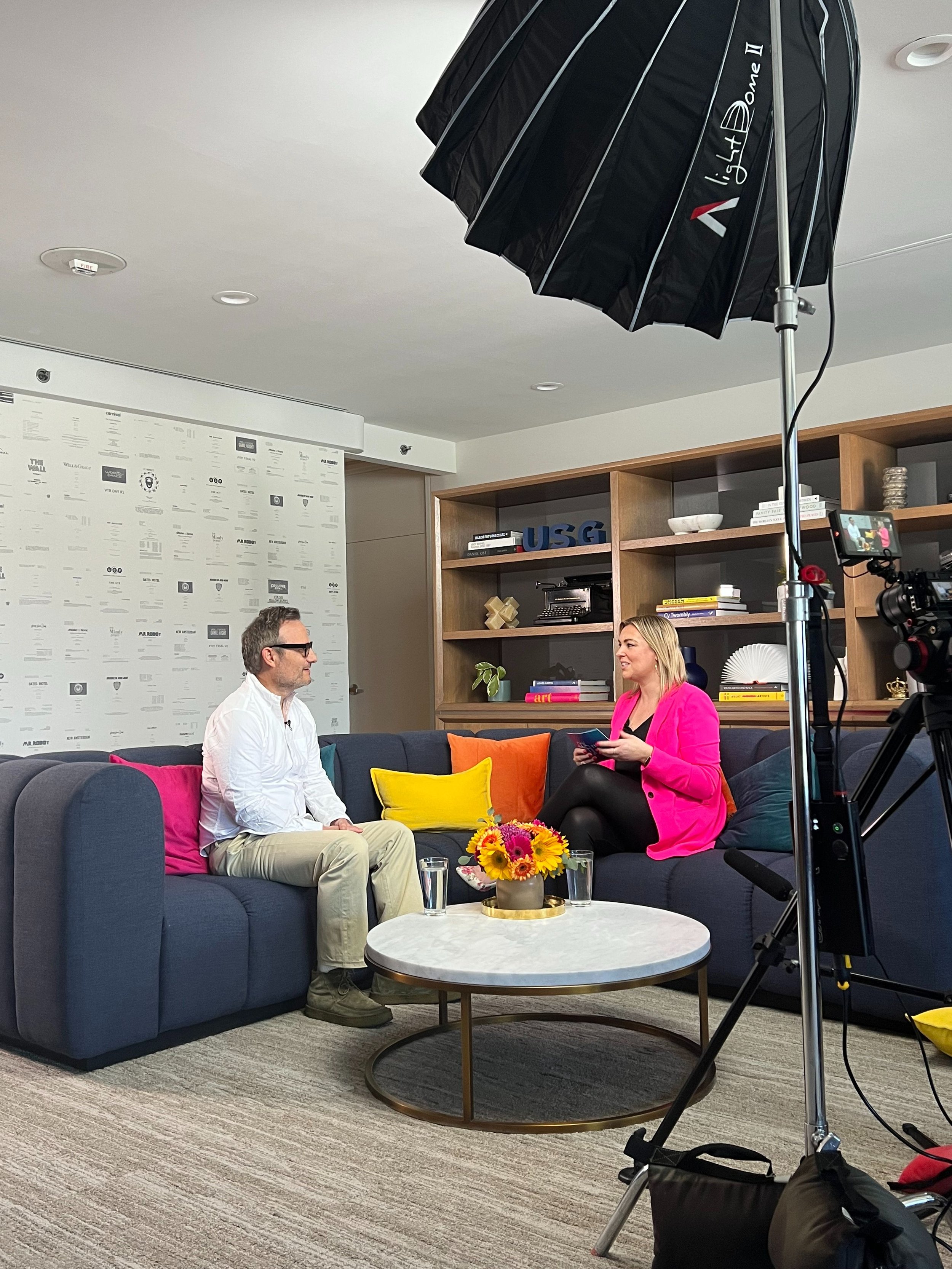 A woman in a bright pink blazer and black pants holding a tablet is sitting on a dark blue sofa with a man in a white shirt and beige pants sitting across from her. They are in a modern, cozy office or lounge area with a wooden bookshelf, colorful pillows, a flower arrangement on a round white coffee table, and filming equipment nearby.