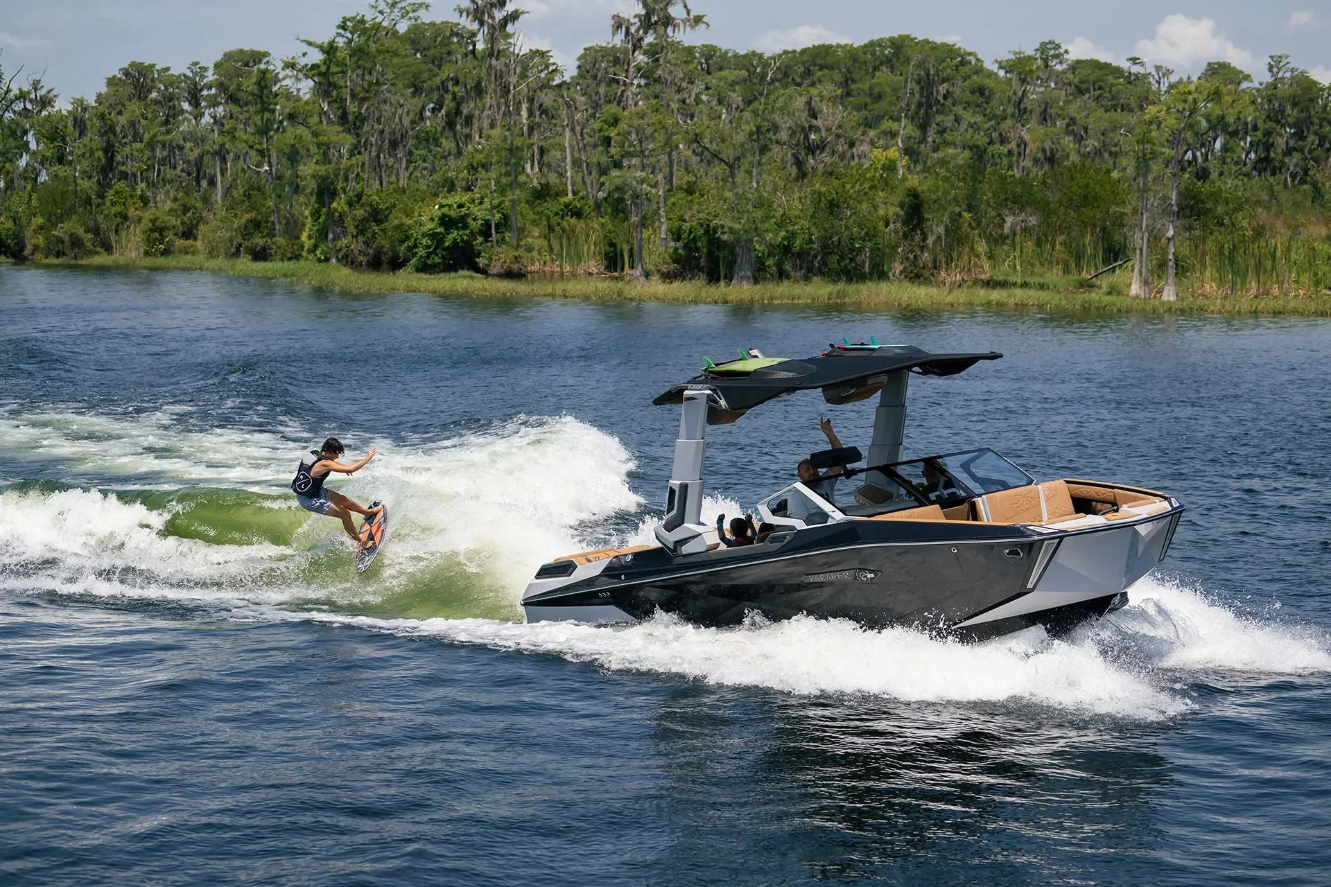 A person wakeboarding behind a motorboat on a lake, with a dense green forest in the background.