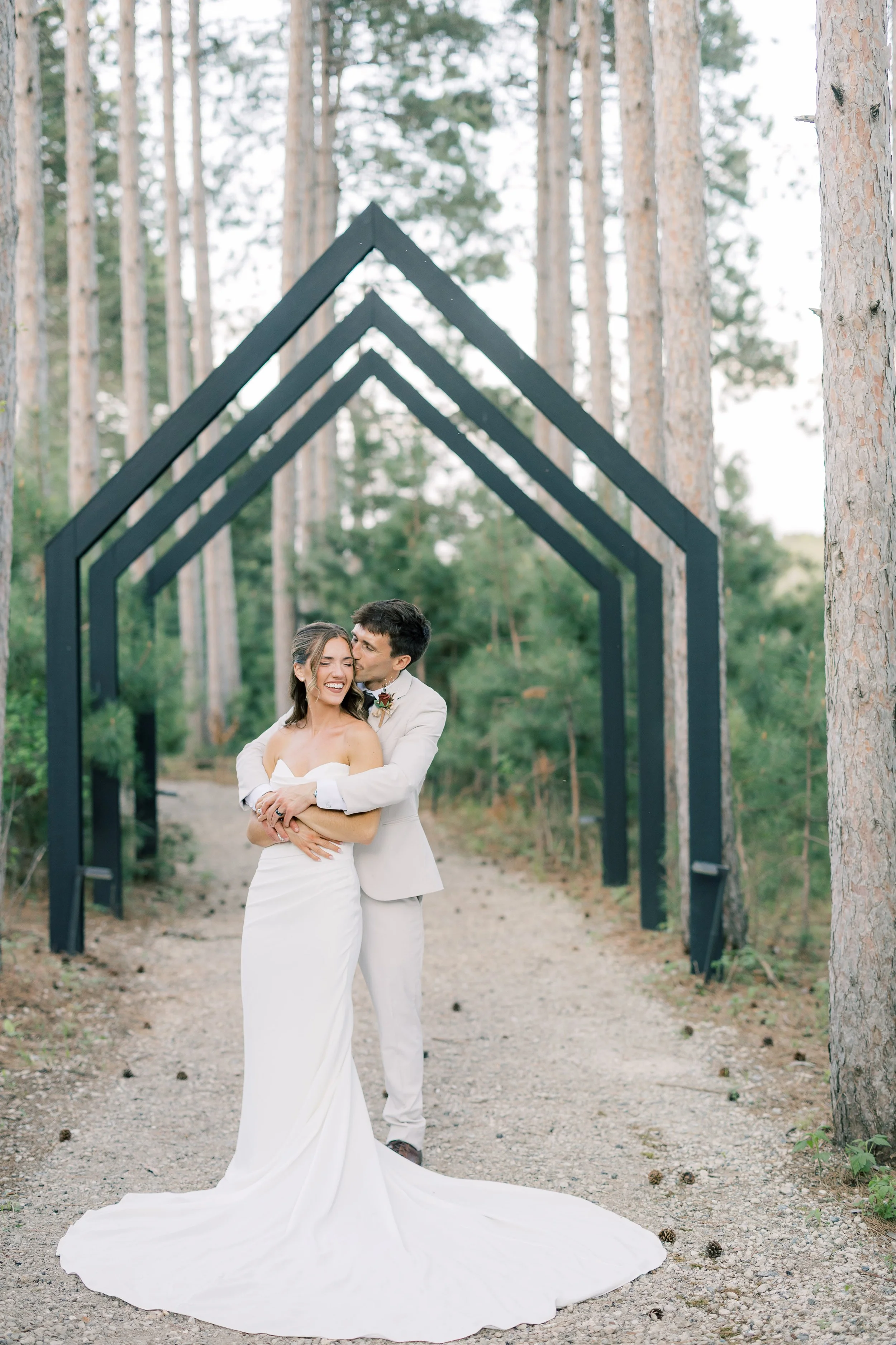 A newlywed couple dressed in white, standing in a wooded outdoor setting with tall pine trees in the background, under a black geometric arch.