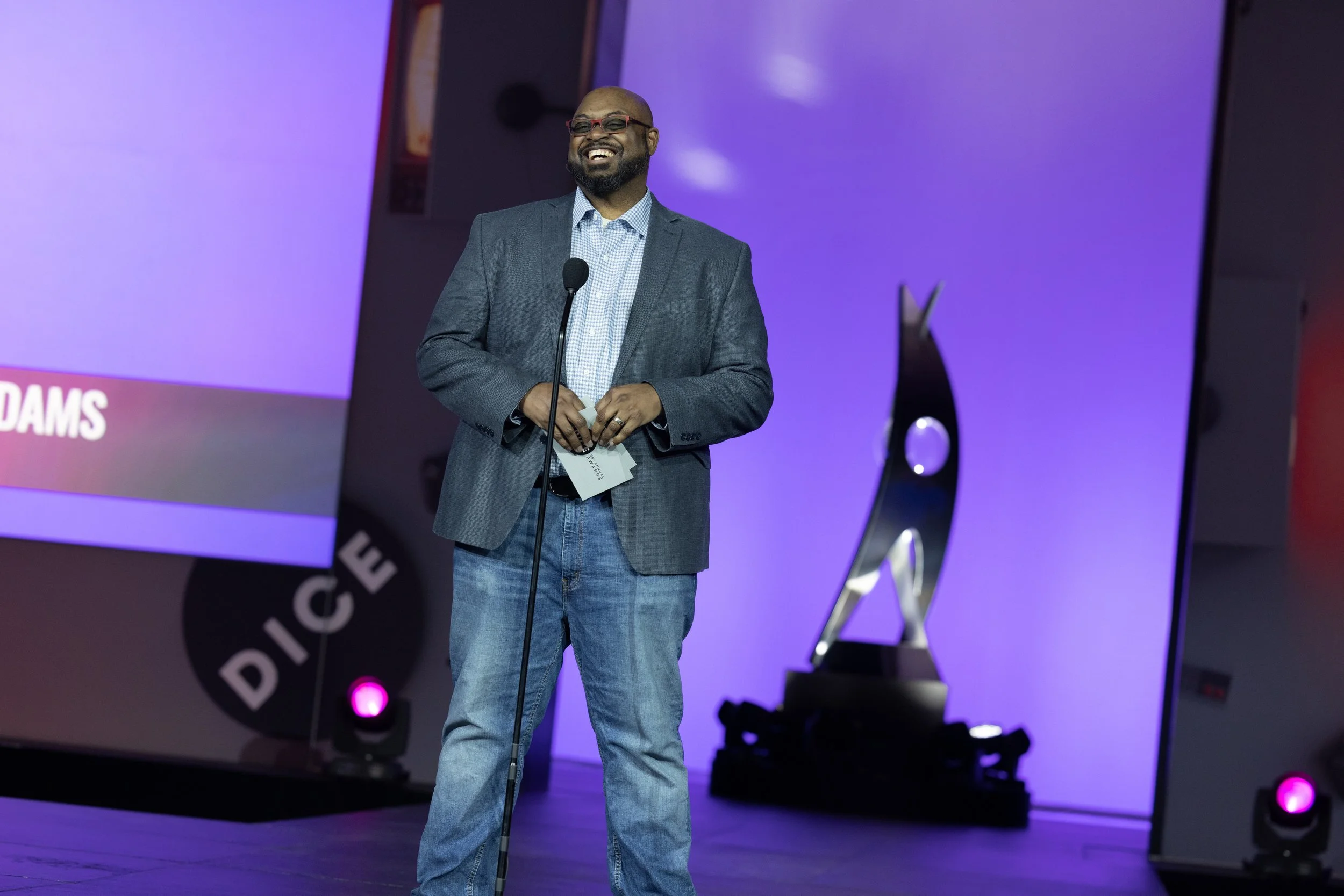 A man in a gray blazer, blue shirt, and jeans is smiling on stage, holding a microphone, with a large screen and stage lights in the background.