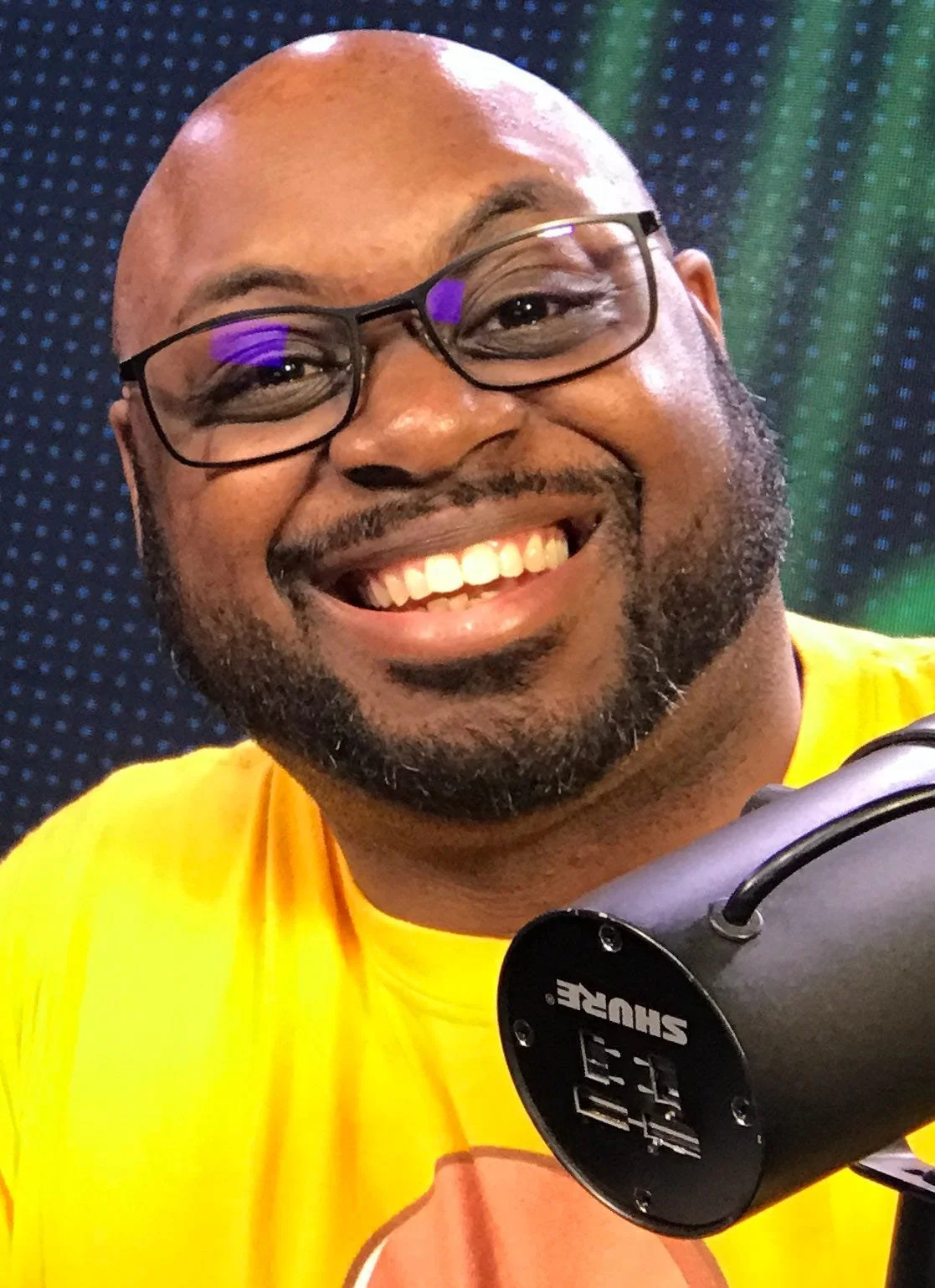 A smiling man wearing glasses and a yellow shirt, holding a device with multiple USB ports, in front of a colorful digital background.