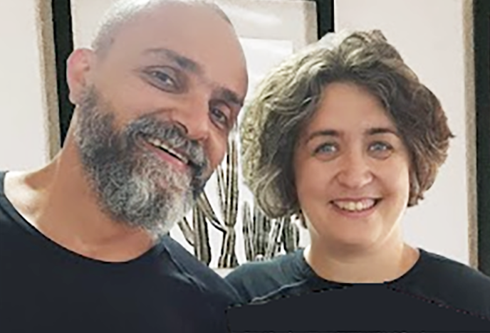A middle-aged man with a beard and a woman with curly hair, both smiling, standing inside a room with framed artwork and decorative vases in the background.