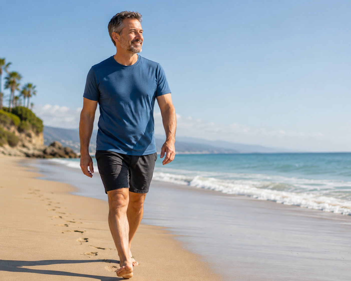 A middle-aged man with gray hair and a beard walking barefoot along the shoreline at the beach, smiling with the ocean and blue sky in the background.