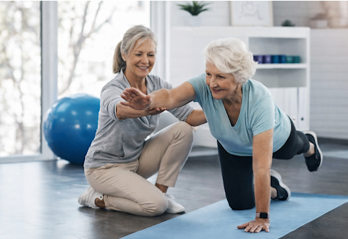 Elderly woman doing a plank exercise with assistance from a younger woman in a bright fitness room.