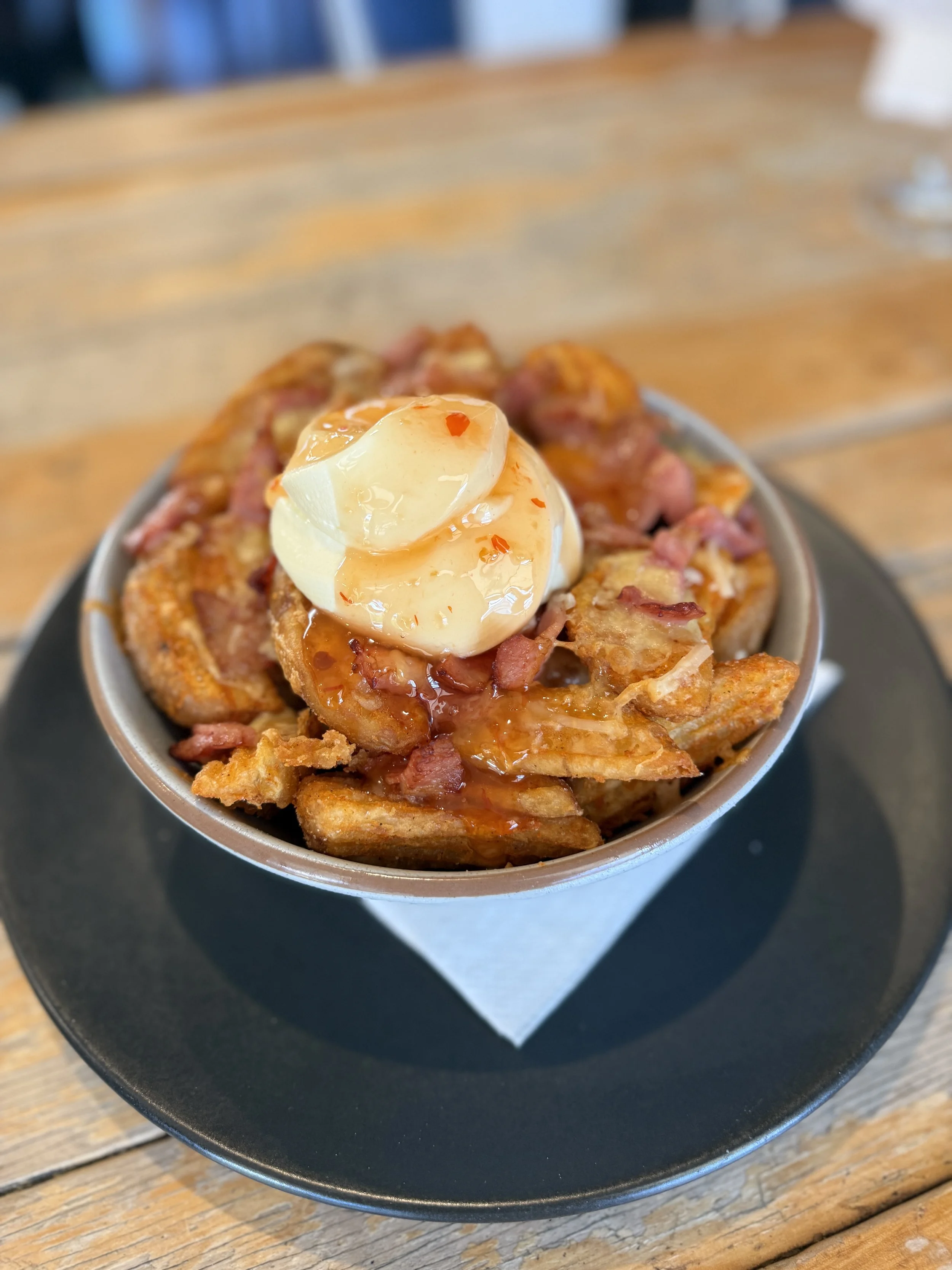A bowl of loaded tater tots topped with cheddar cheese, diced ham, a poached egg, and drizzled with hot sauce, placed on a black plate on a wooden table.