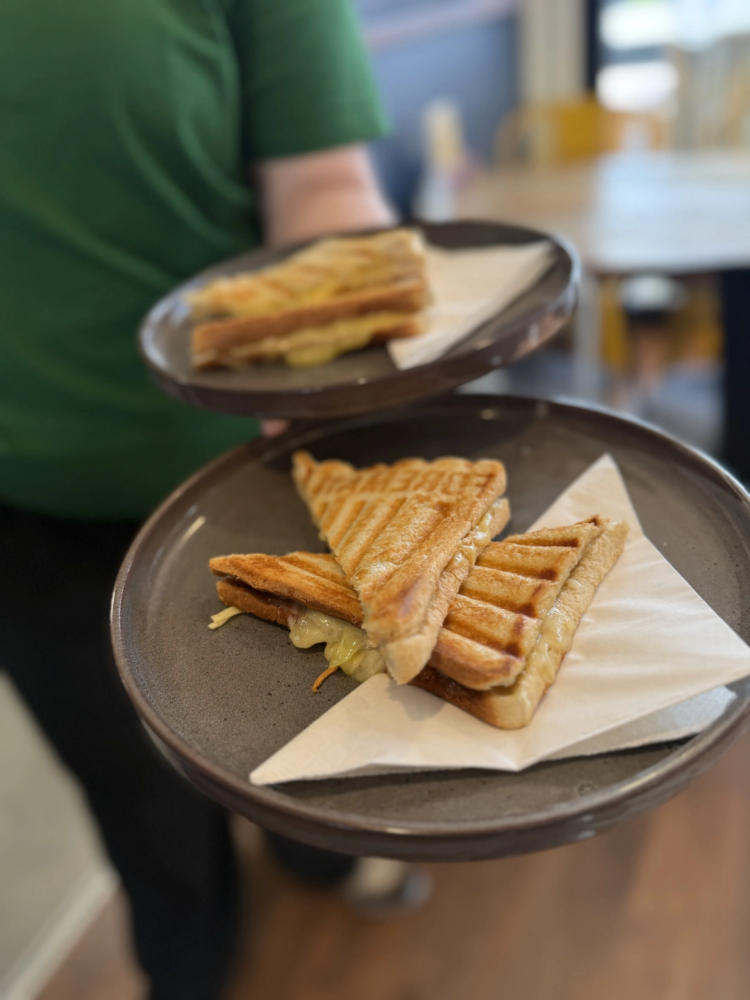 Close-up of a grilled sandwich with melted cheese and lettuce on a brown plate, held by a person wearing a green shirt, in a restaurant.