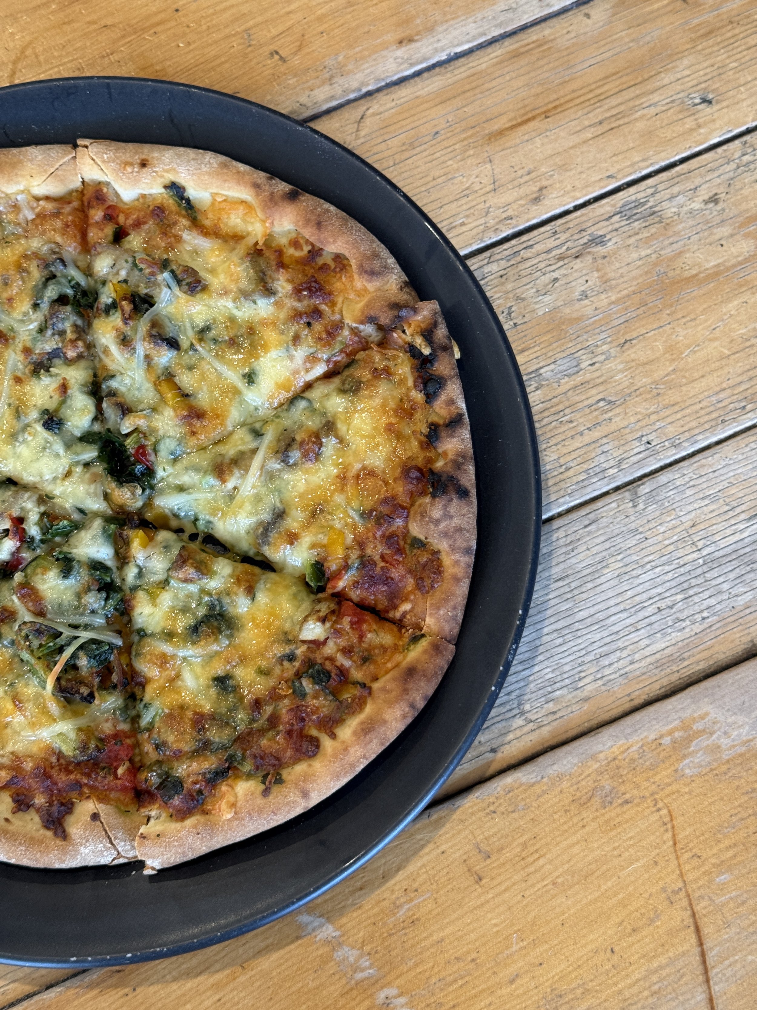 A partially visible pizza with cheese, vegetables, and possibly meat on a black plate on a wooden table.