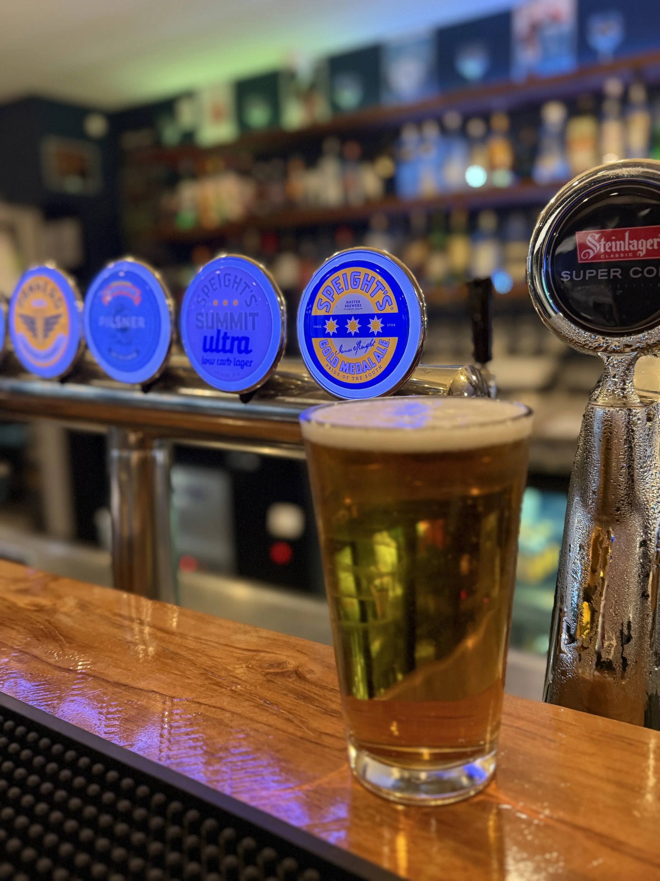 A pint of beer on a bar counter with blue tap handles labeled Speight's beers in a bar setting.
