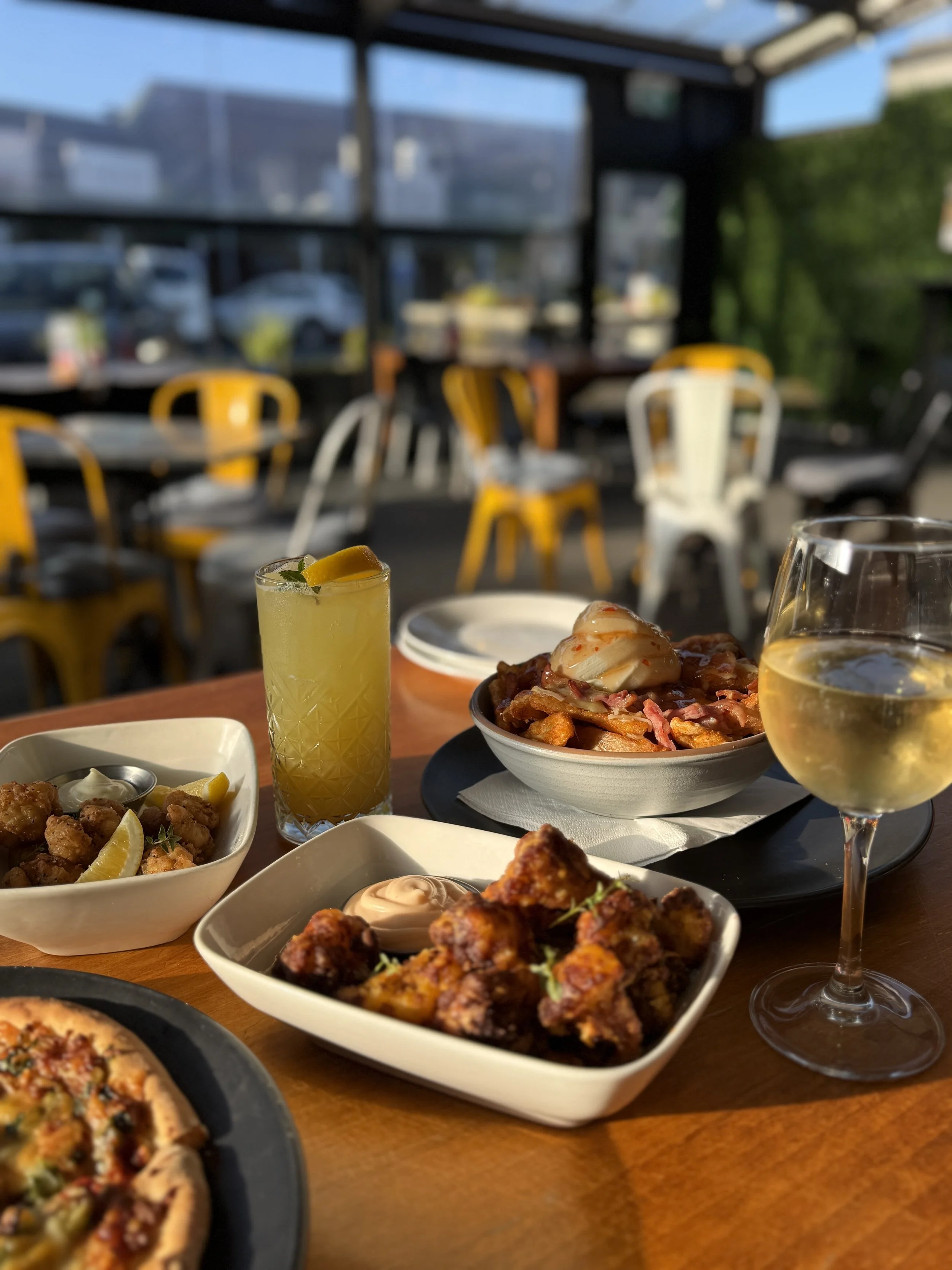 A table with various foods including fried chicken, pizza, and salads, along with drinks such as a lemon beverage and white wine, in a restaurant with large windows and yellow and white chairs.