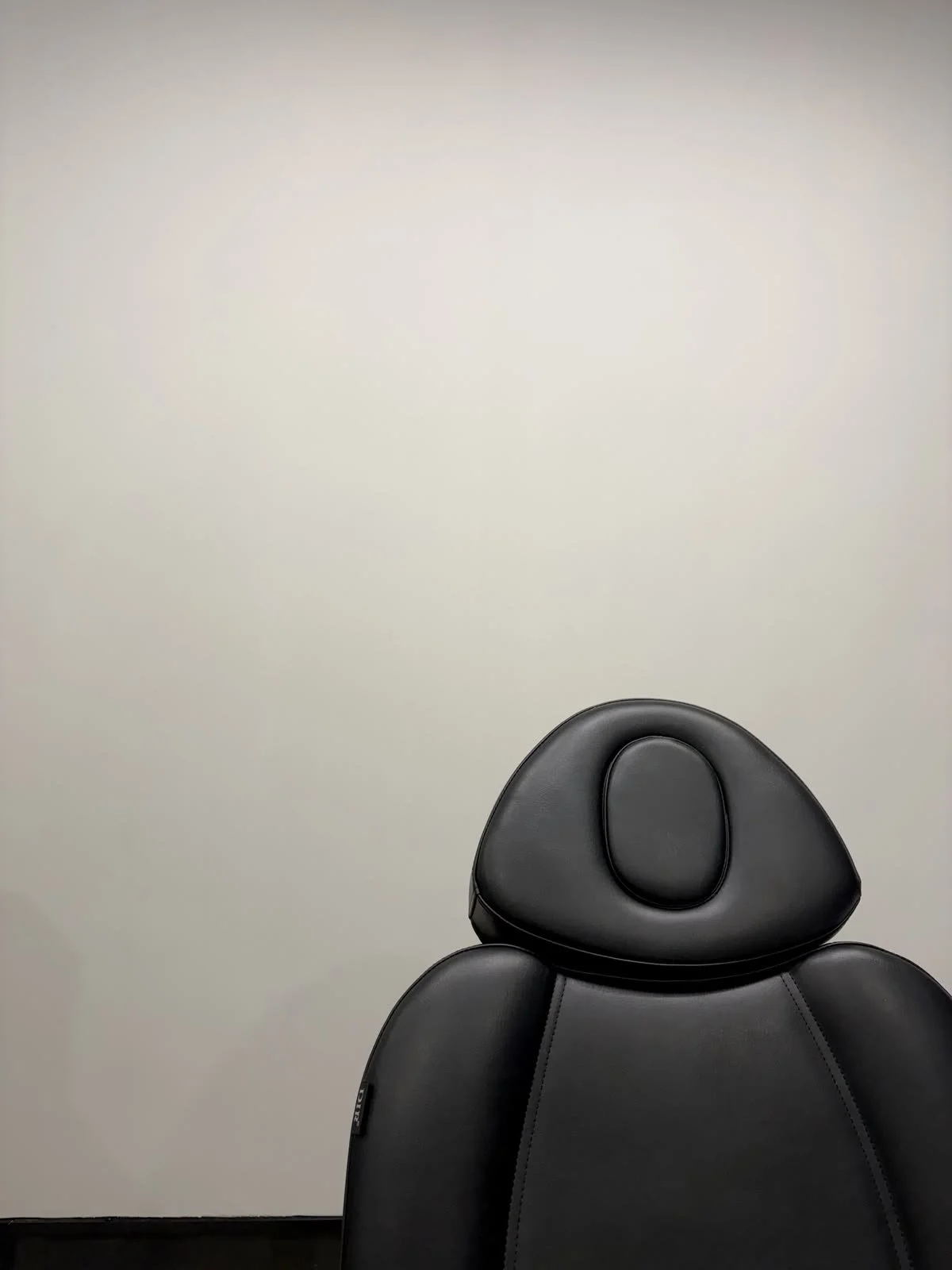 Part of a black leather or faux leather seat with a headrest, placed against a plain white wall.