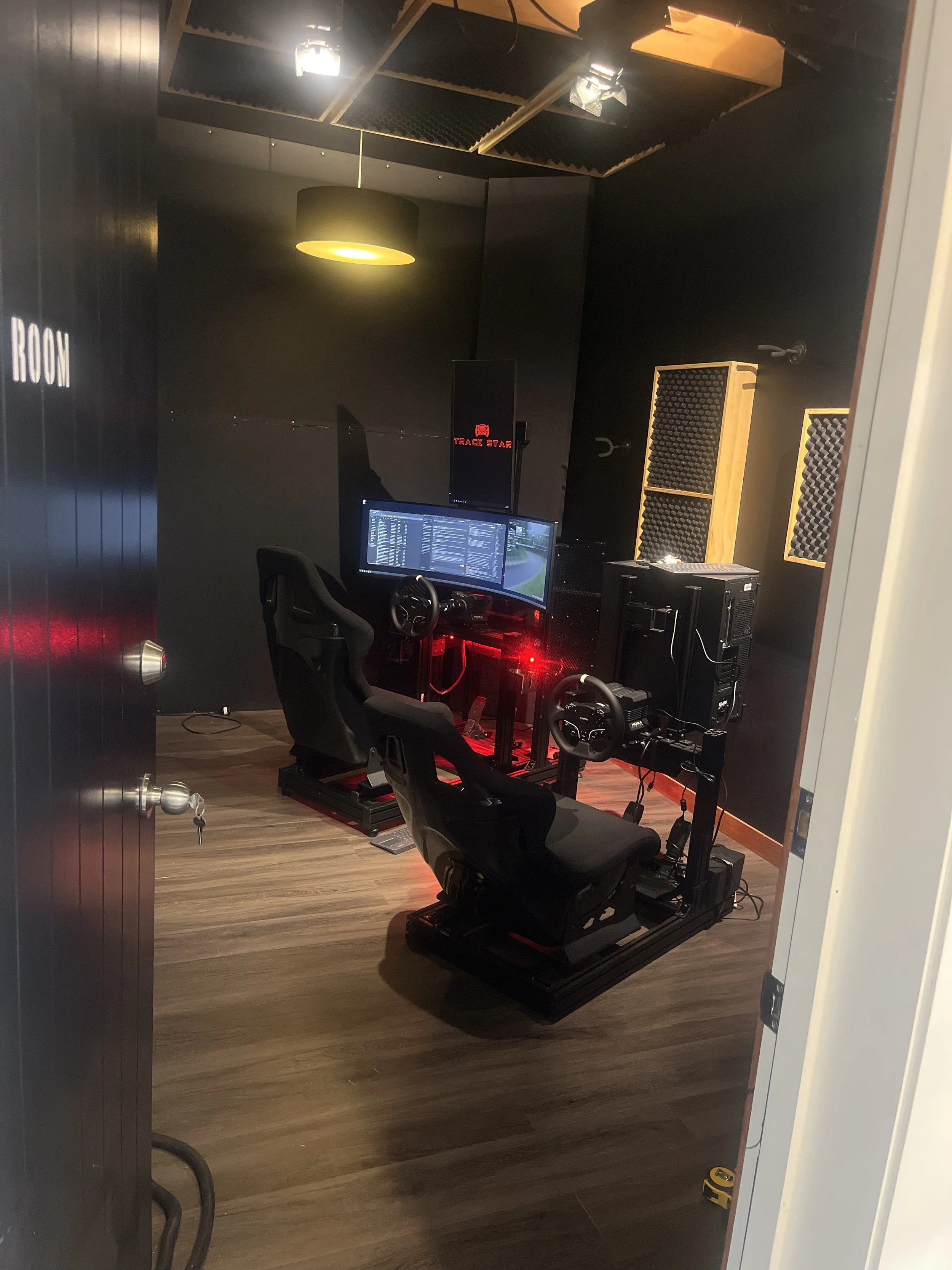 A room with black walls and ceiling, equipped with a racing simulator setup including a racing seat, steering wheel, pedals, and three computer monitors displaying software. The room has specialized soundproofing panels on the walls and a yellow pendant light and fluorescent ceiling lights.