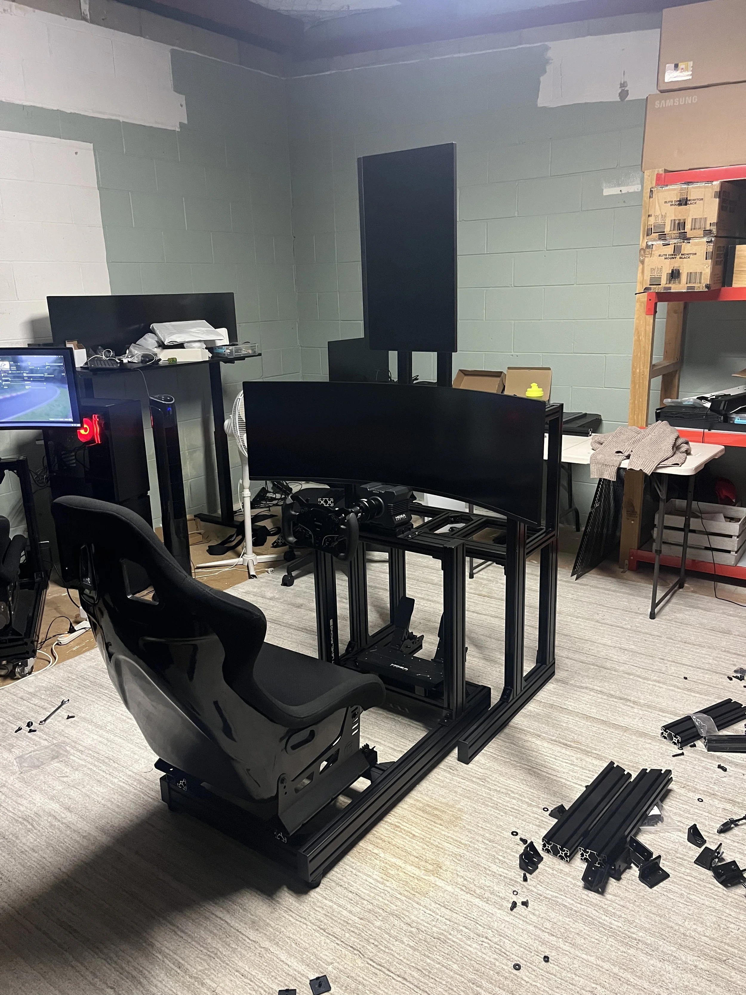 A room with moving computer monitors, a chair, parts and tools scattered on the floor, and a partially assembled computer desk frame.
