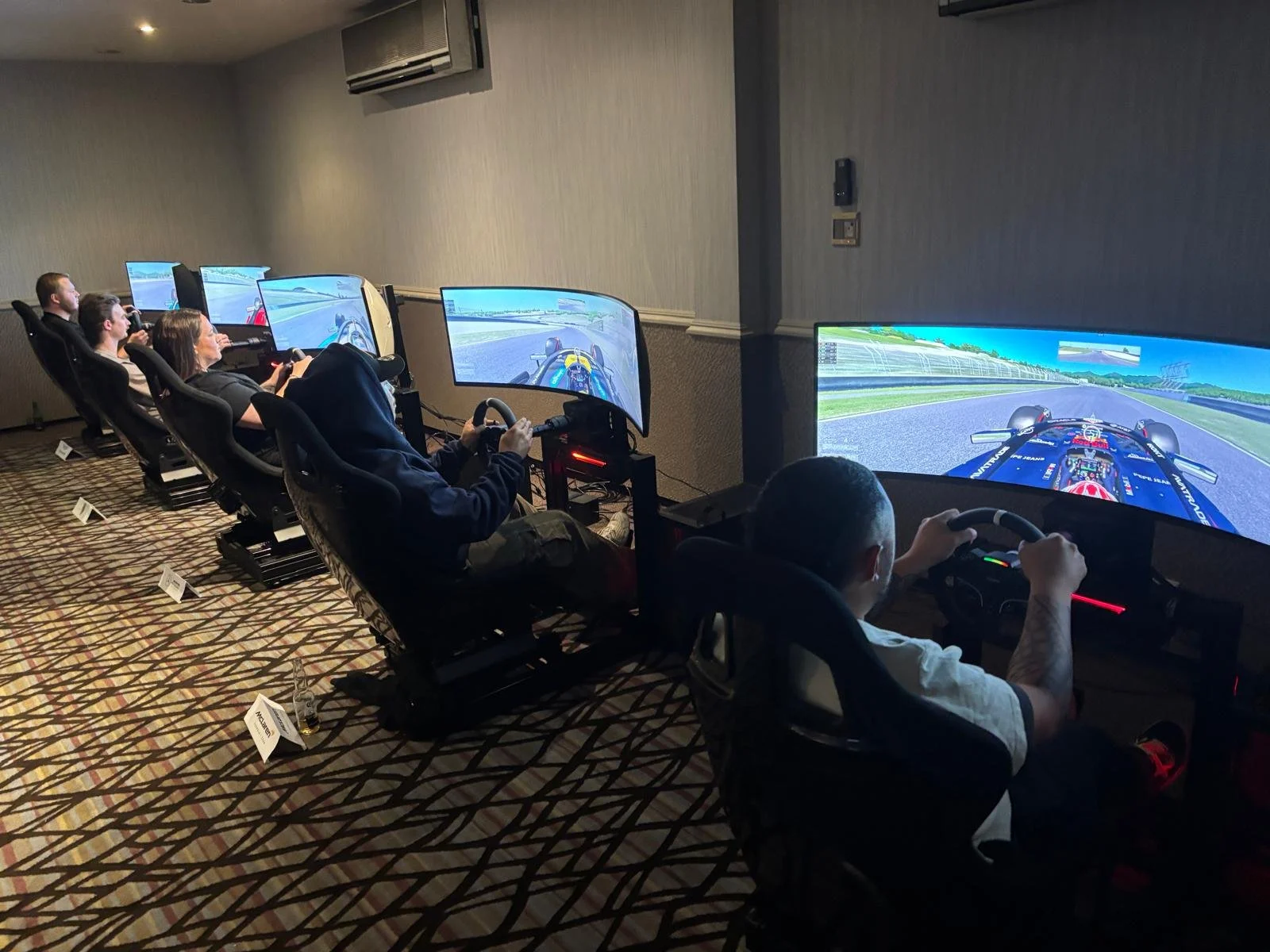 People seated in racing simulator chairs with large curved screens in a room, driving a simulated race car on a virtual racetrack.