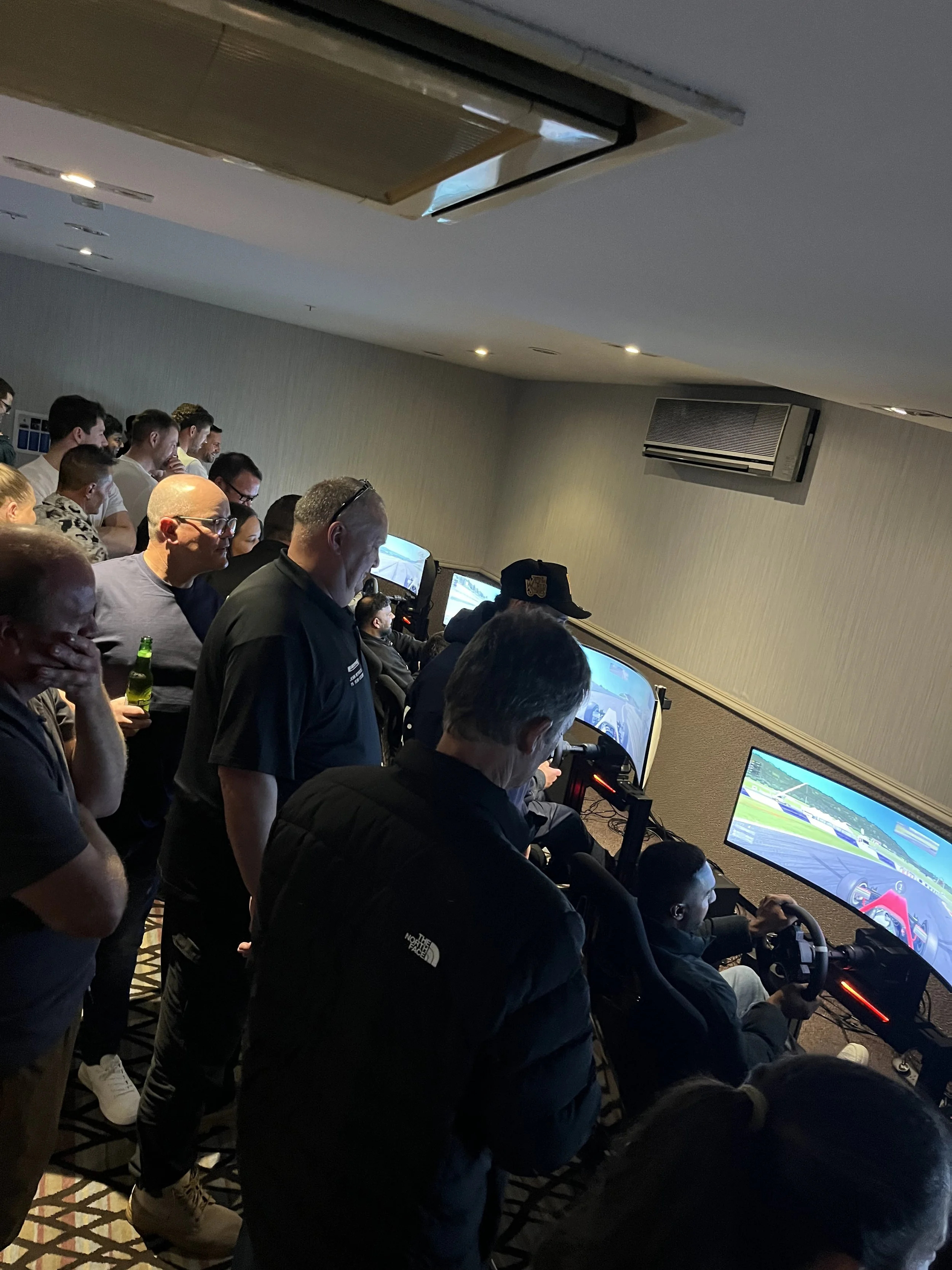 A group of people are gathered in a room, playing racing simulation games on multiple screens with steering wheels and pedals. The room has a low ceiling, beige walls, and ceiling-mounted air conditioning units.