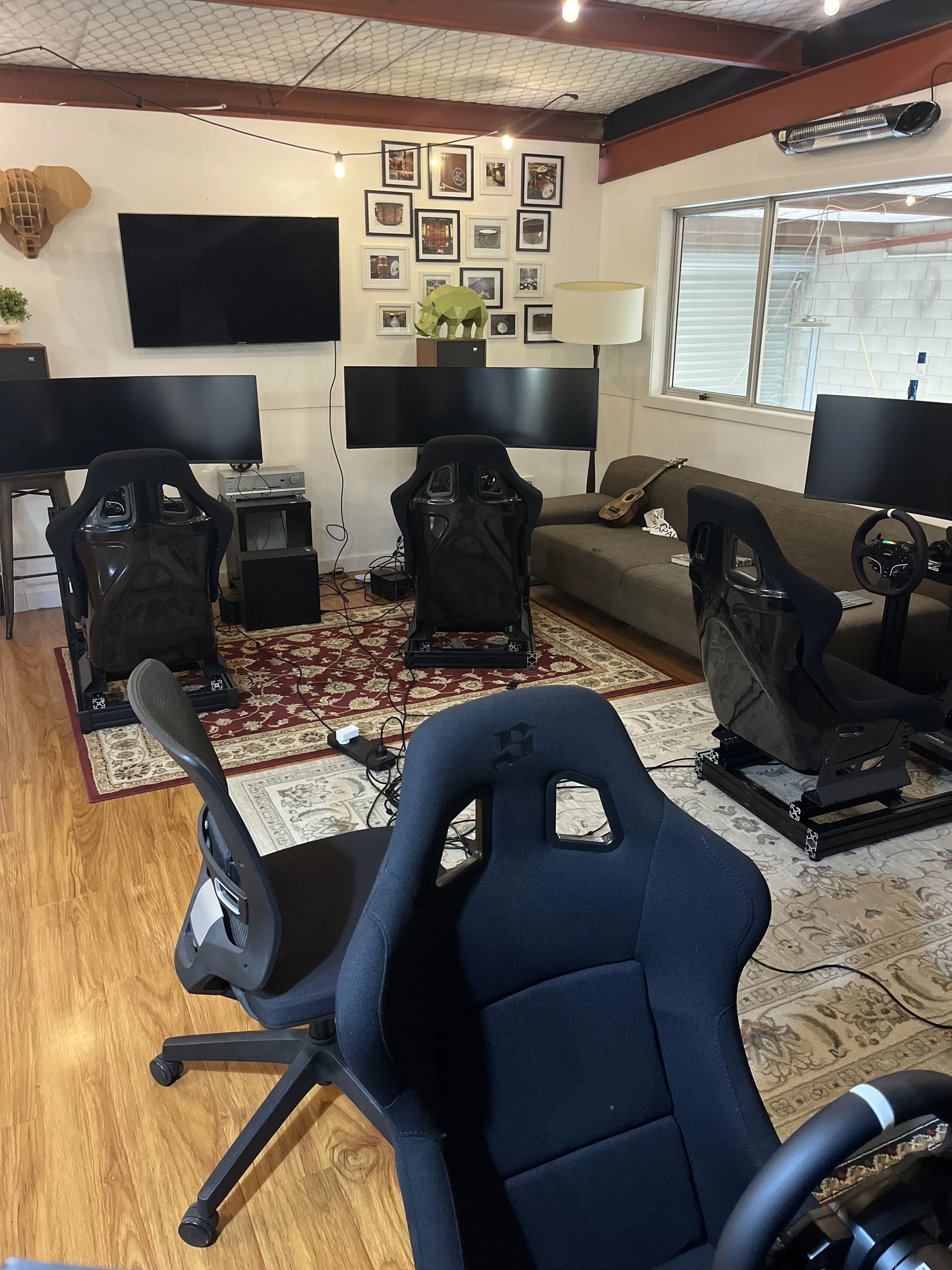A room set up with multiple gaming chairs, several computer monitors, a TV on the wall, and gaming accessories. There is a couch with a small guitar and gaming controller on it. The room has a wooden floor, a rug, and a window with blinds.