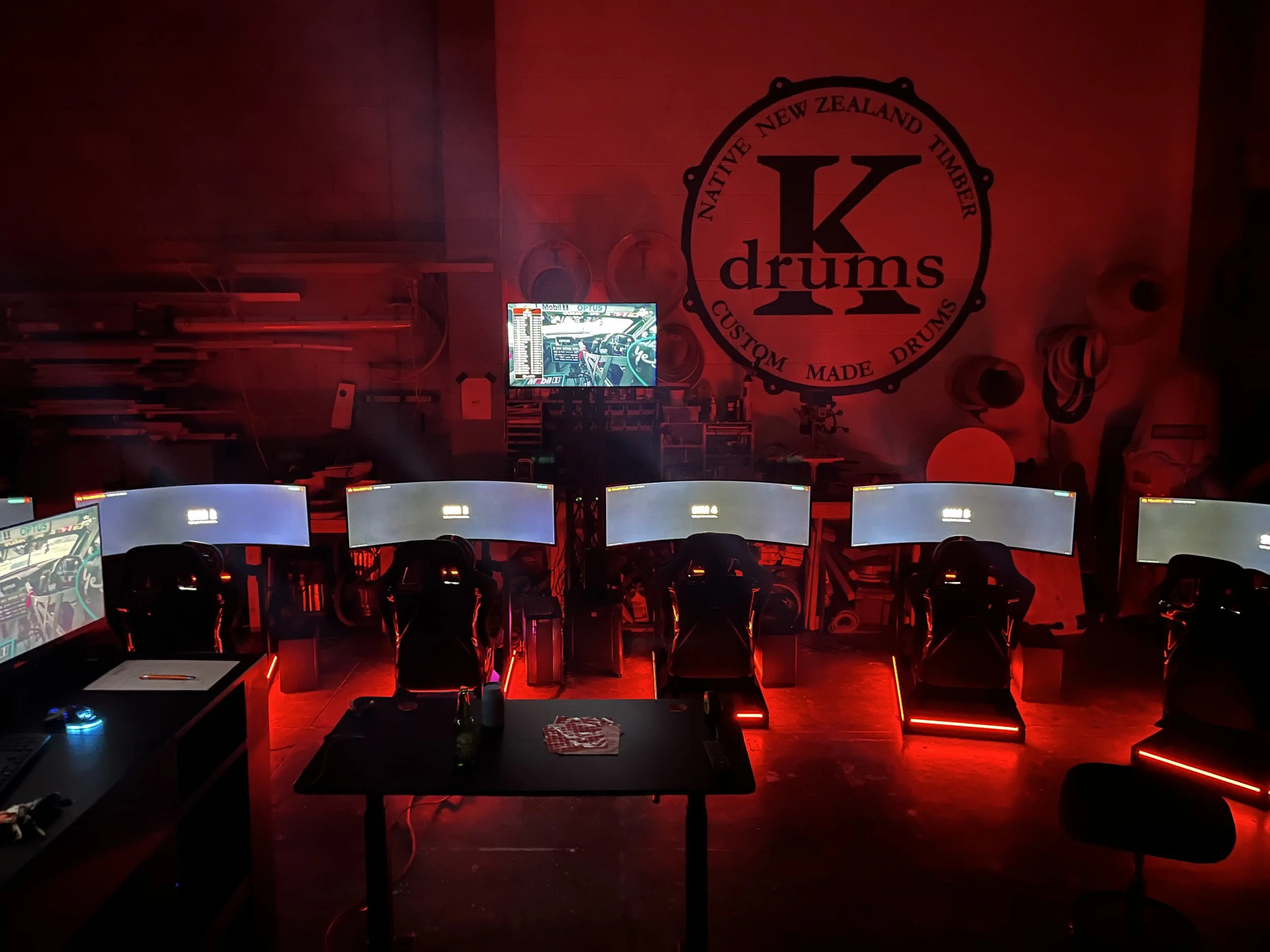 An arcade or gaming room with multiple computer stations, red lighting, a wall with a logo reading 'Ng drums, New Zealand, Timber, Custom, Made Drums', and a television showing a racing game.