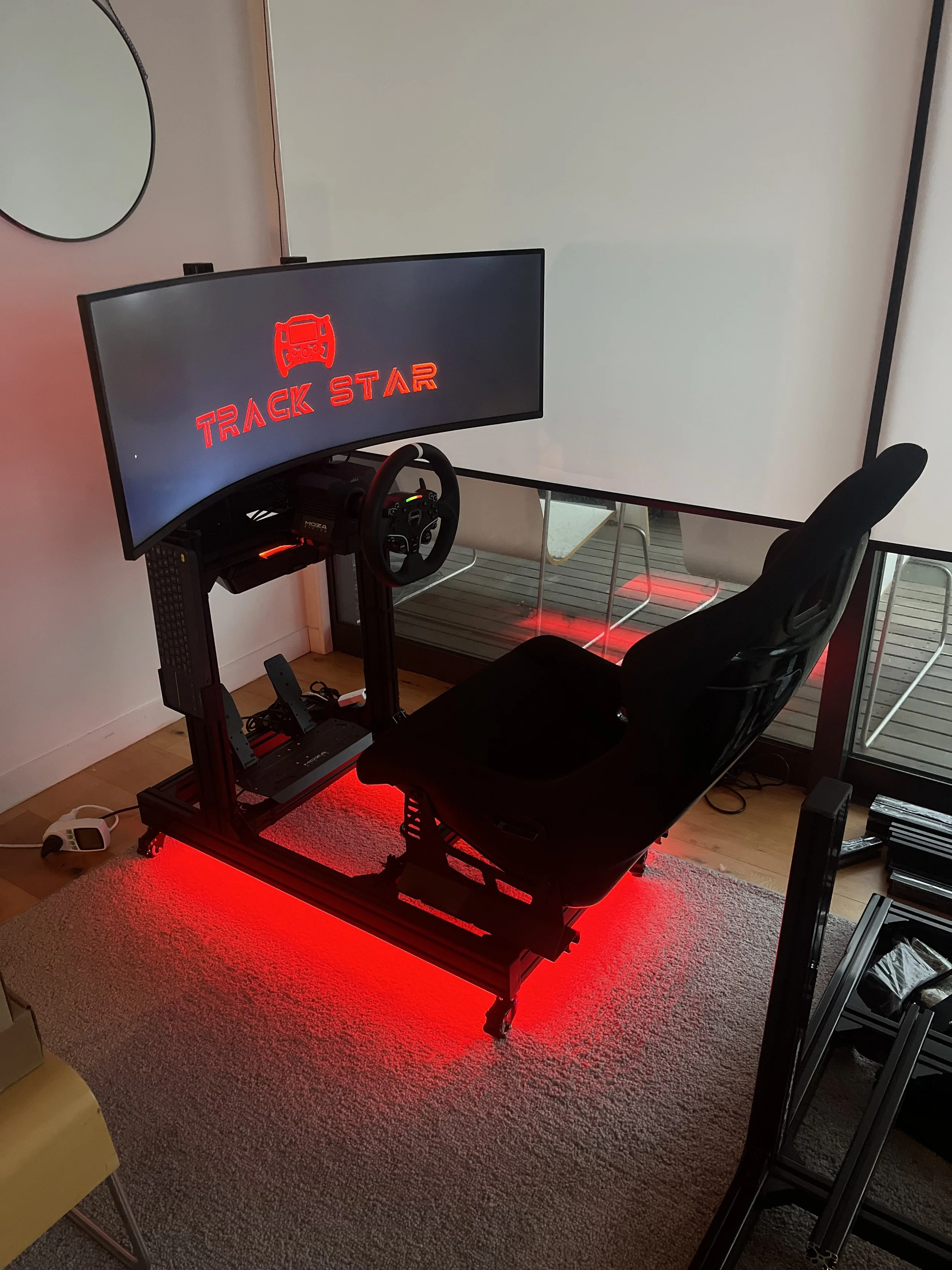 A racing simulator setup with a curved monitor displaying the words 'TRACK STAR' and a steering wheel, sitting on a desk with red LED lighting underneath. There is a black gaming chair in front of the setup, and a white wall and balcony with outdoor furniture in the background.