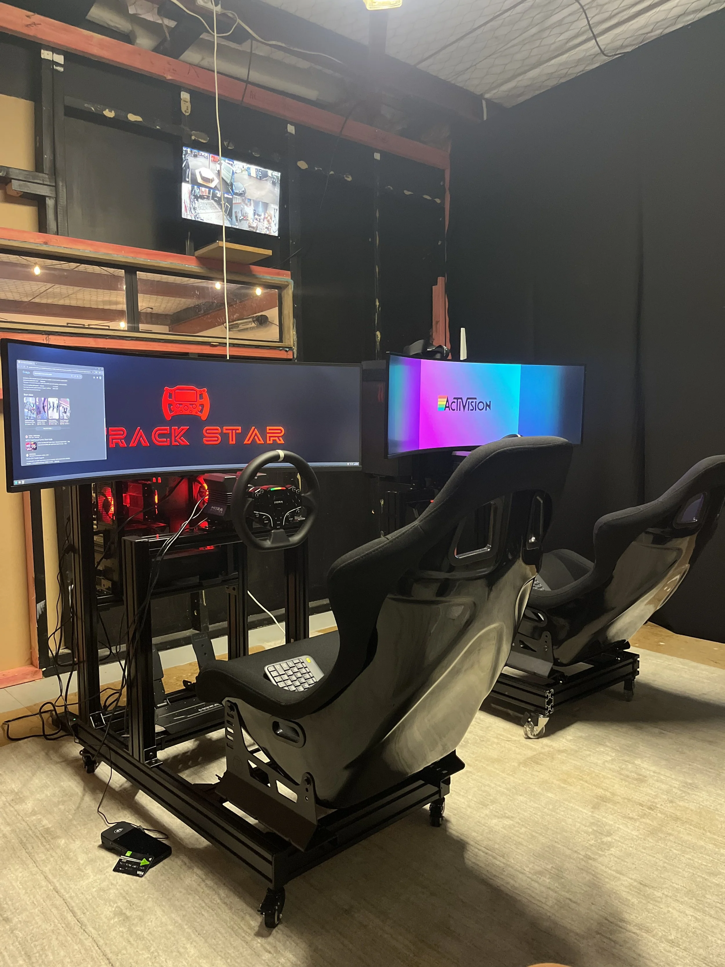 A racing simulator setup with a black racing seat, three monitors, a steering wheel, and pedals on a wooden floor. The monitors display a logo and colorful background, and there is a large mirror on the wall behind the setup.