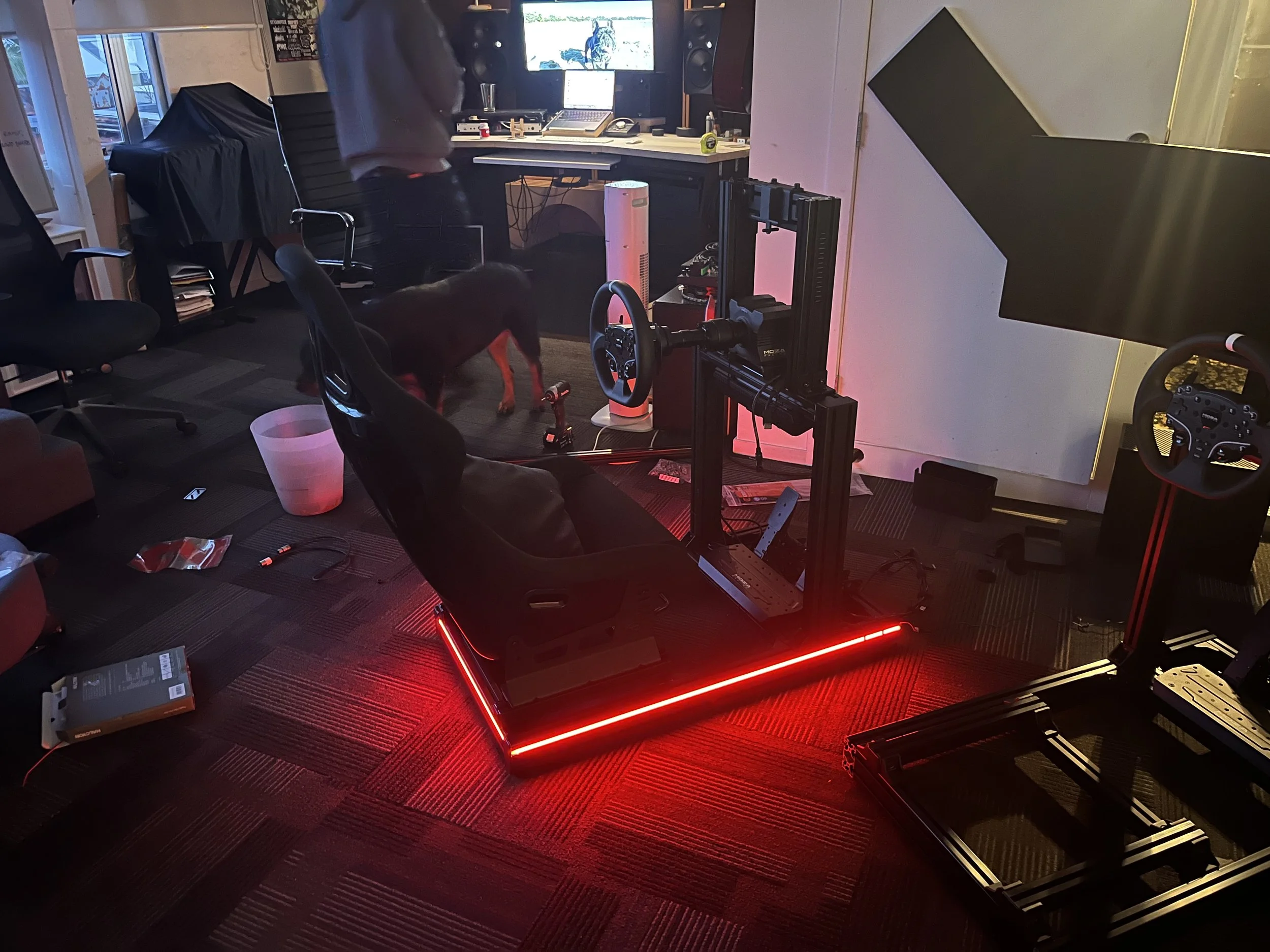 A person working in a home office with computer equipment, a large monitor on the wall, a 3D printer, a black dog, and various office supplies. The room has a dark floor with red LED lighting around a 3D printer and a window showing the outdoors.