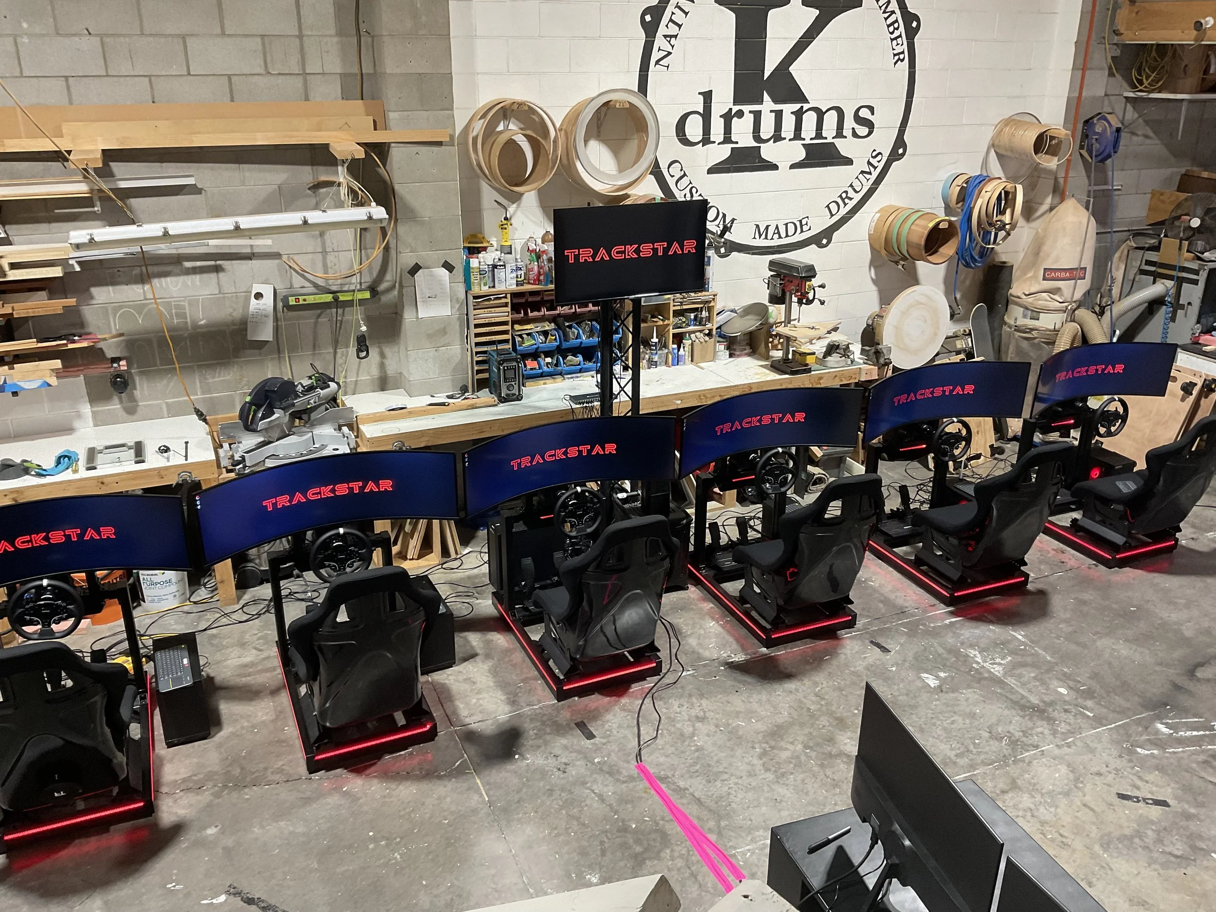 A workshop with multiple gaming simulators featuring racing car seats and large curved screens showing the Trackstar logo, with woodworking tools and supplies on the workbench and walls.