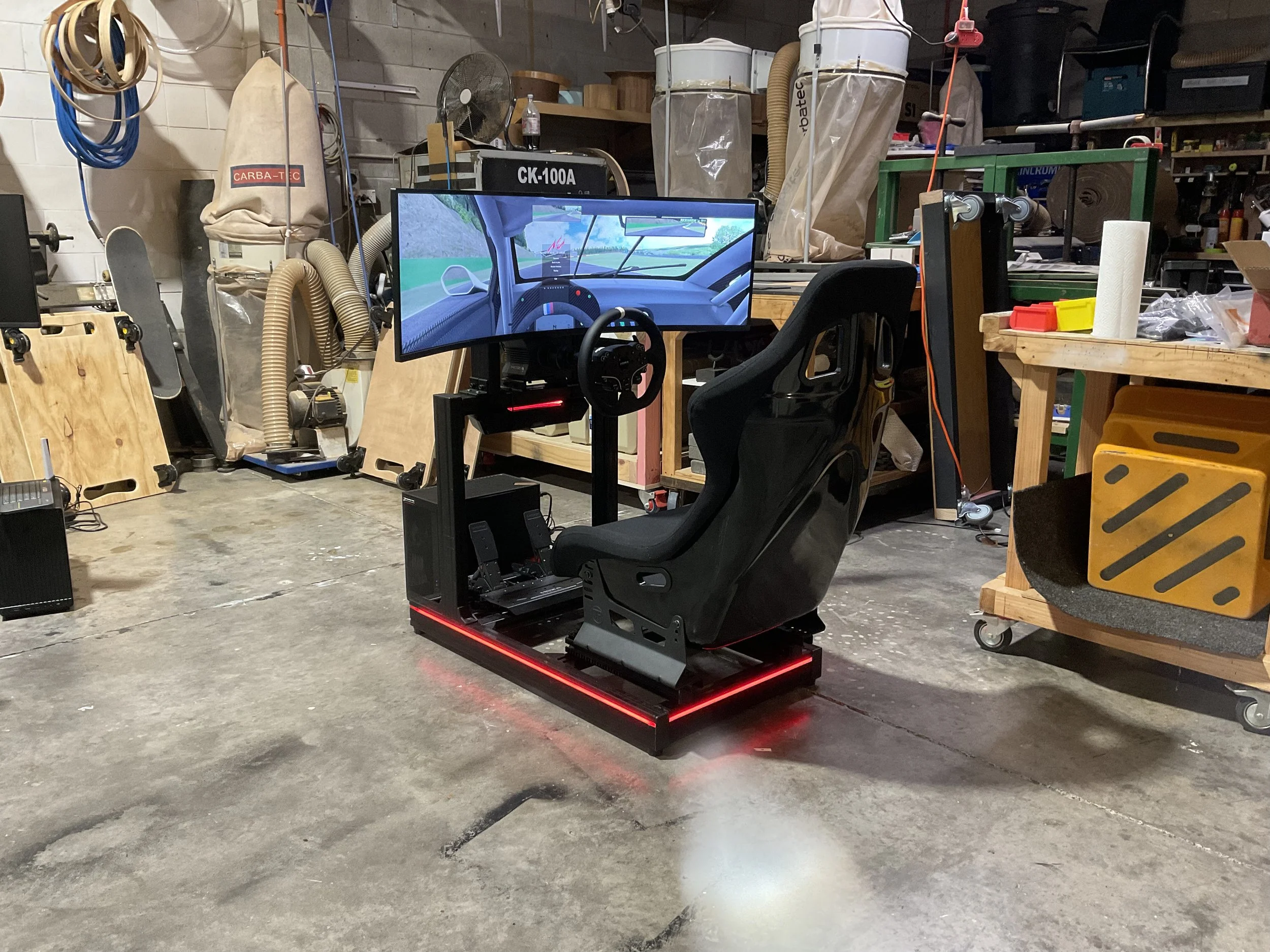 A racing simulator setup with a large curved screen displaying a driving game, a racing seat, and pedals, situated in a cluttered workshop or garage with tools and supplies.