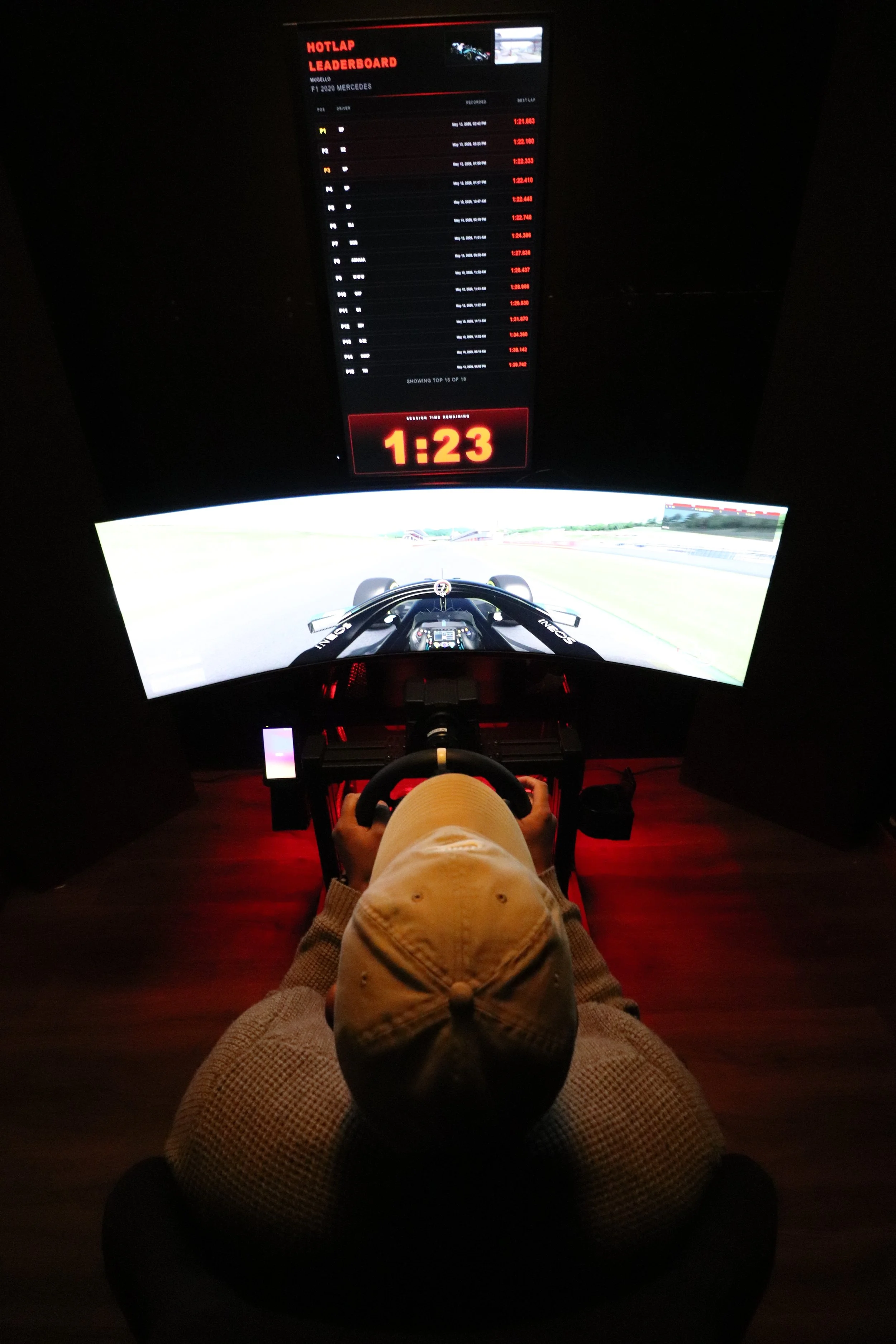 A person sitting in a racing simulator setup with a steering wheel and pedal controls. The screen shows a virtual race track with a car on the road. Above the screen, a digital leaderboard displays race times, and a large digital clock shows 1:23.