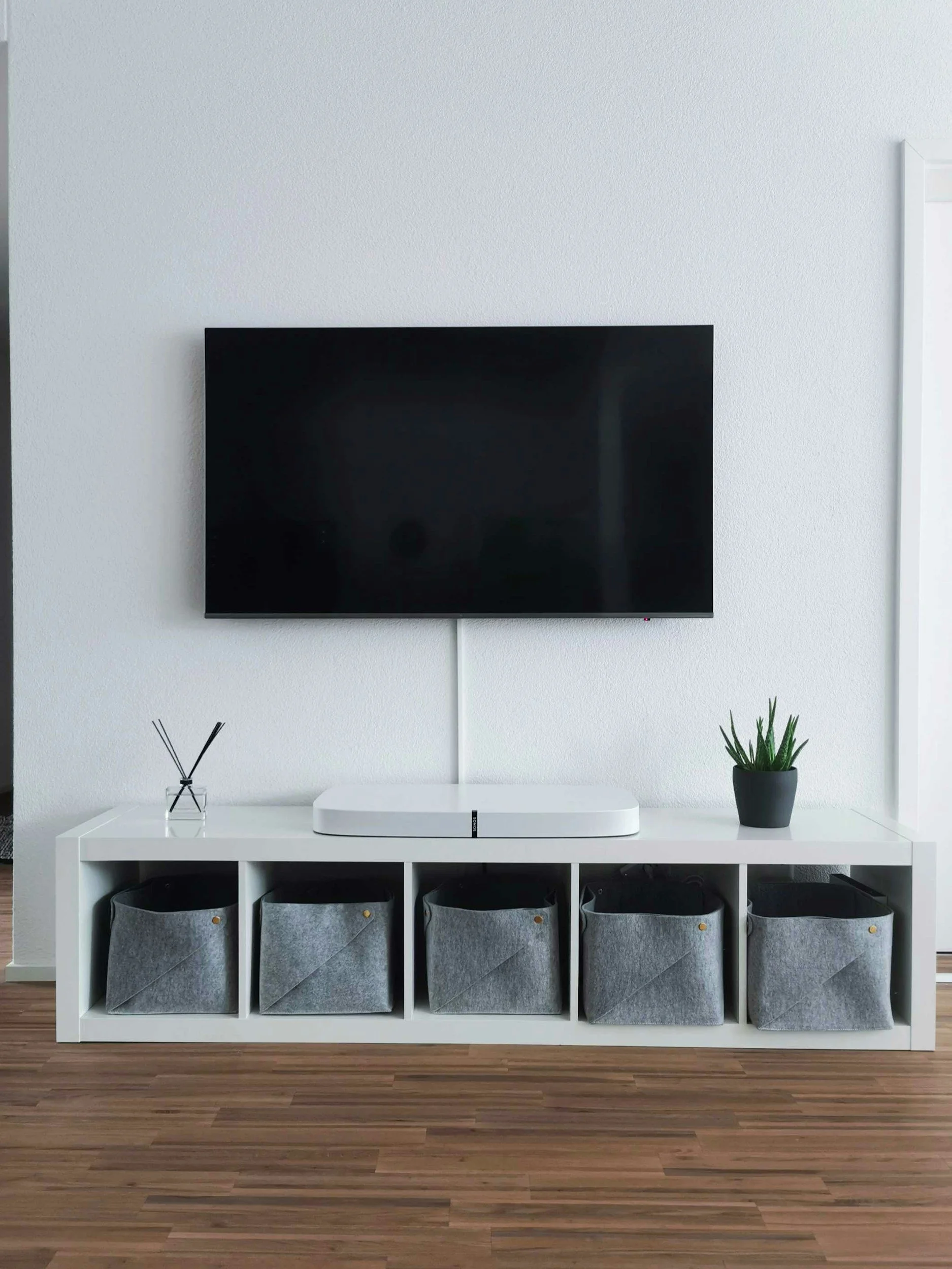 A modern living room setup with a white console table against a white textured wall. On top of the table is a black flat-screen TV mounted on the wall, a white media device, a small potted green plant, and a black reed diffuser. Under the table are four fabric storage bins in grey, stored in cubbies. The room has wooden flooring.