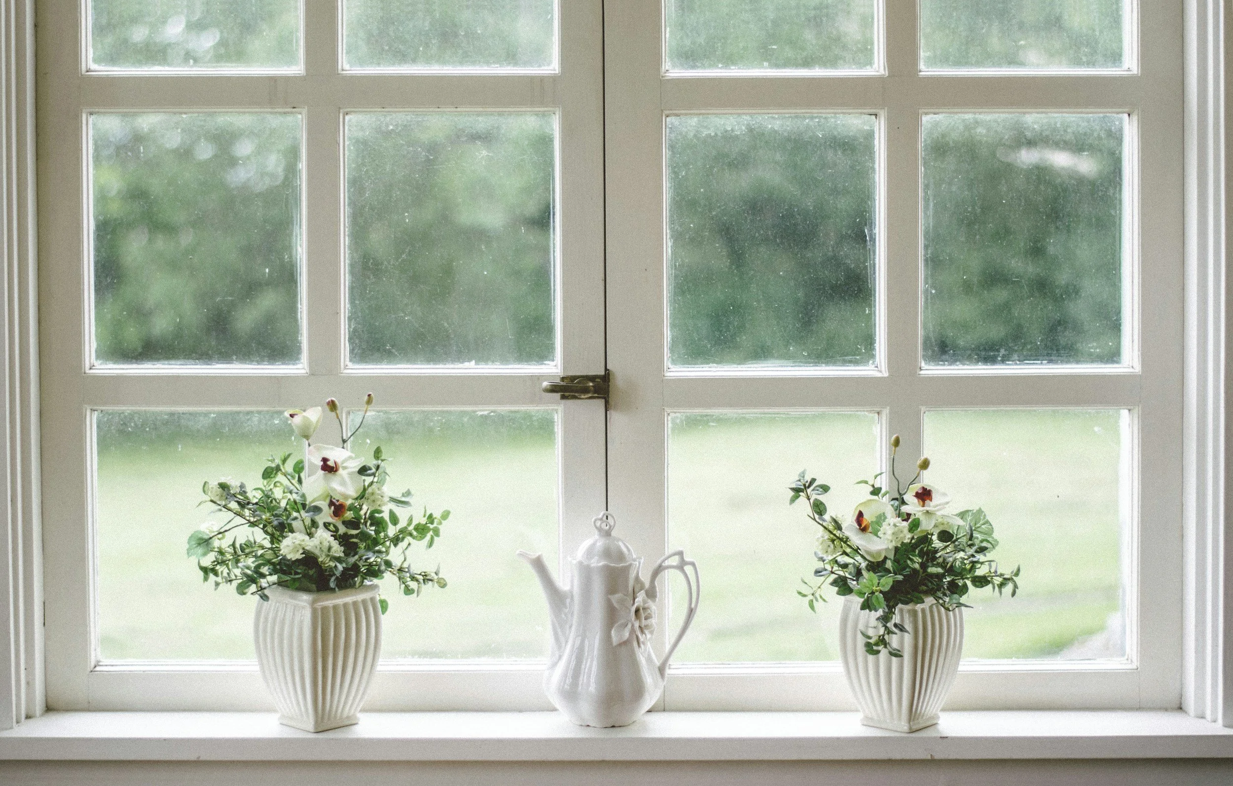 Three white vases with flowers on a window sill with a view of a rainy landscape outside.