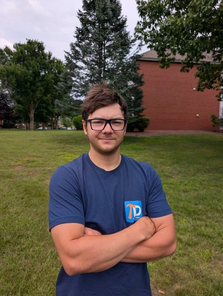 Young man with glasses, crossed arms, wearing a blue T-shirt with a hammer logo, standing on grass in a park with trees and a brick building in the background.
