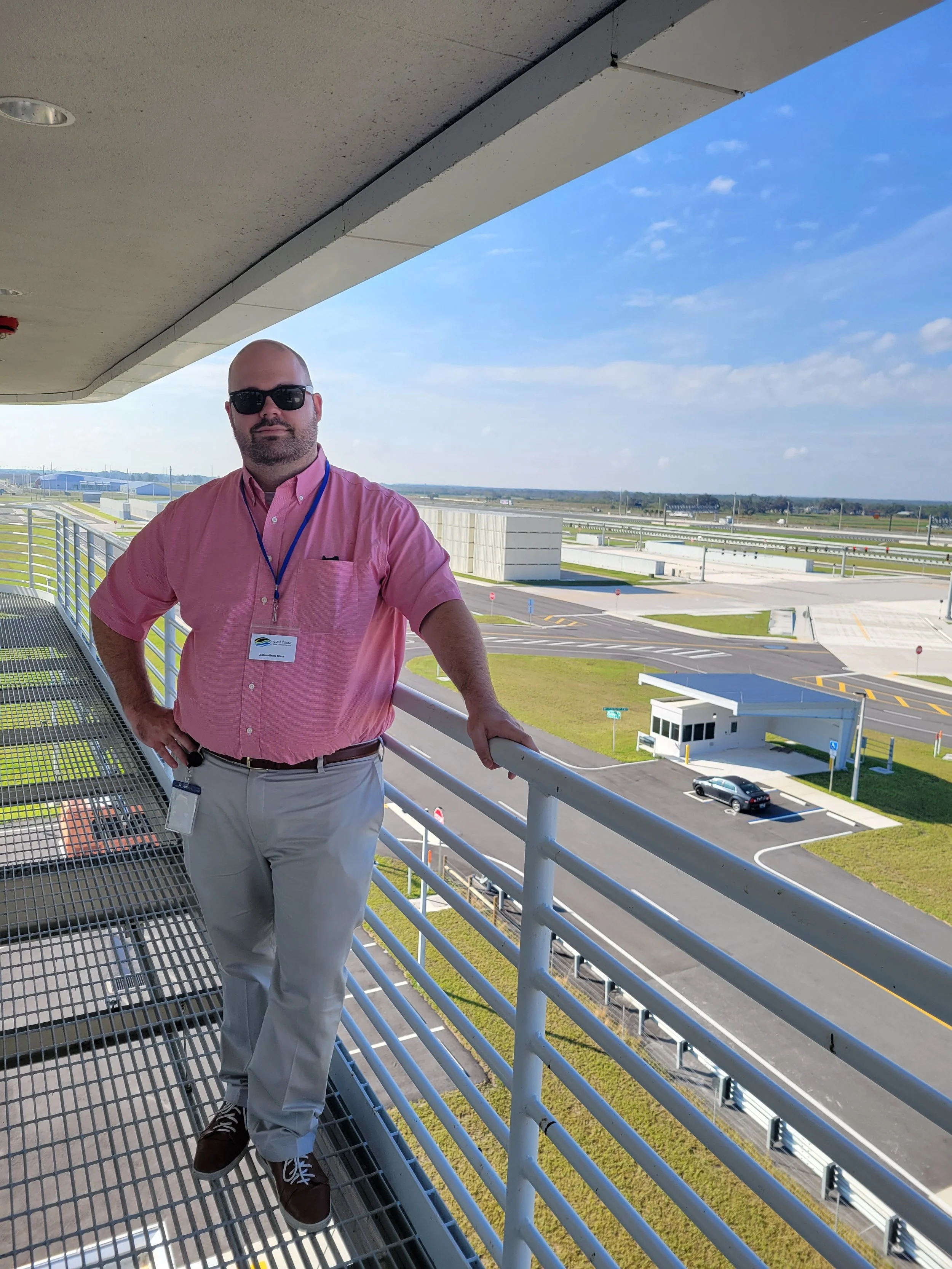 Man wearing sunglasses, pink collared shirt, khaki pants, and a name badge standing on an outdoor balcony of a parking lot or transportation hub, with a view of empty parking spaces, roads, and a flat landscape in the background.