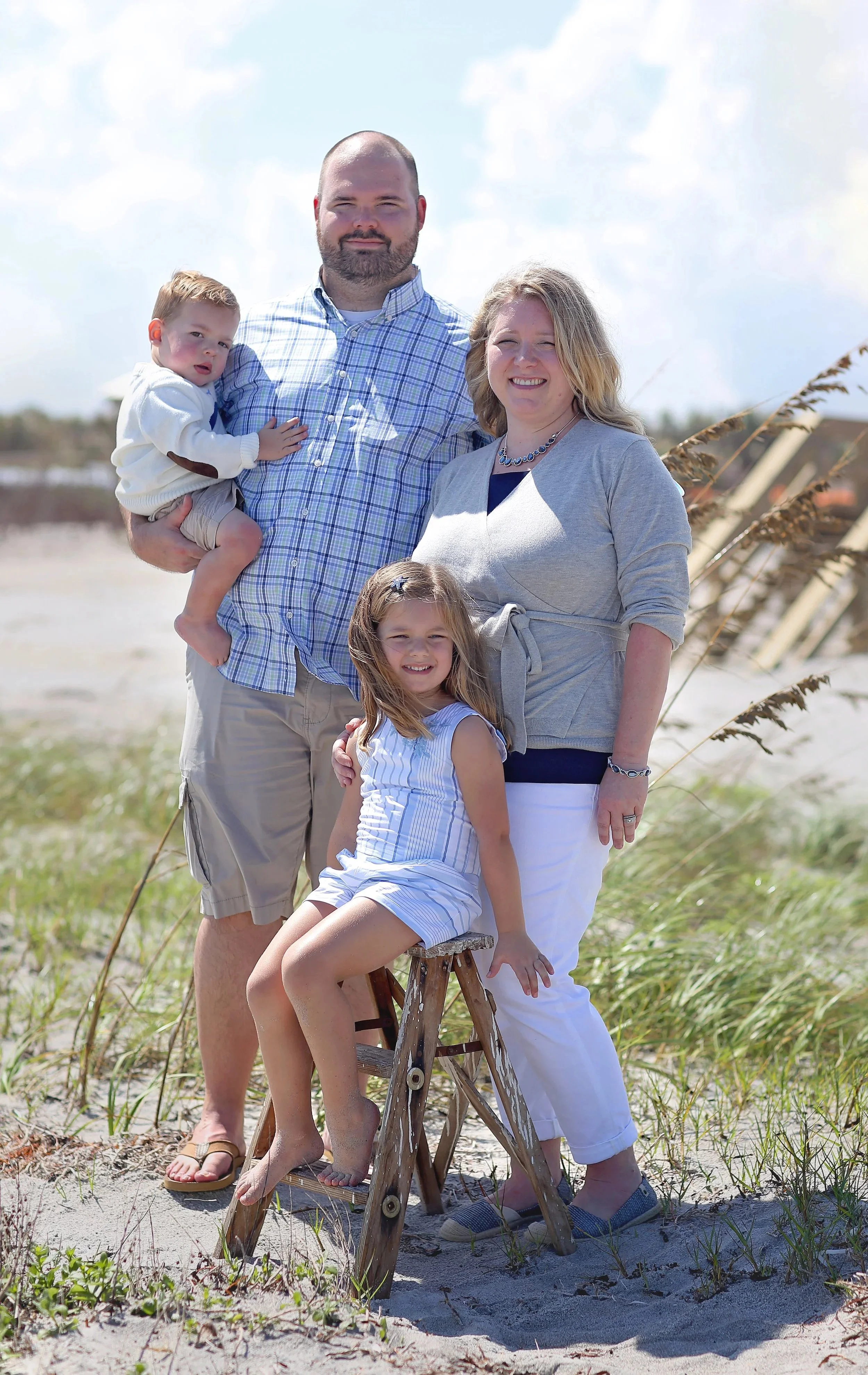 A family of four outdoors on a beach, with tall grasses and a bright sky in the background. The father holds a young boy, and a woman stands next to them with a girl sitting on a small wooden stepladder in front. All are smiling or looking at the camera.
