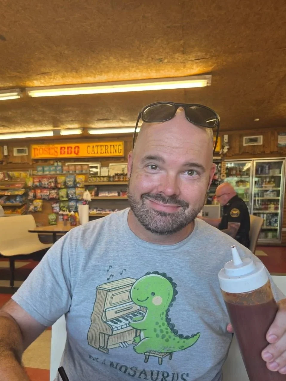 Man with sunglasses on his head, holding a bottle of barbecue sauce, sitting in a casual dining area with a "Rose's BBQ Catering" sign in the background. He is wearing a gray T-shirt with a cartoon dinosaur playing the piano.