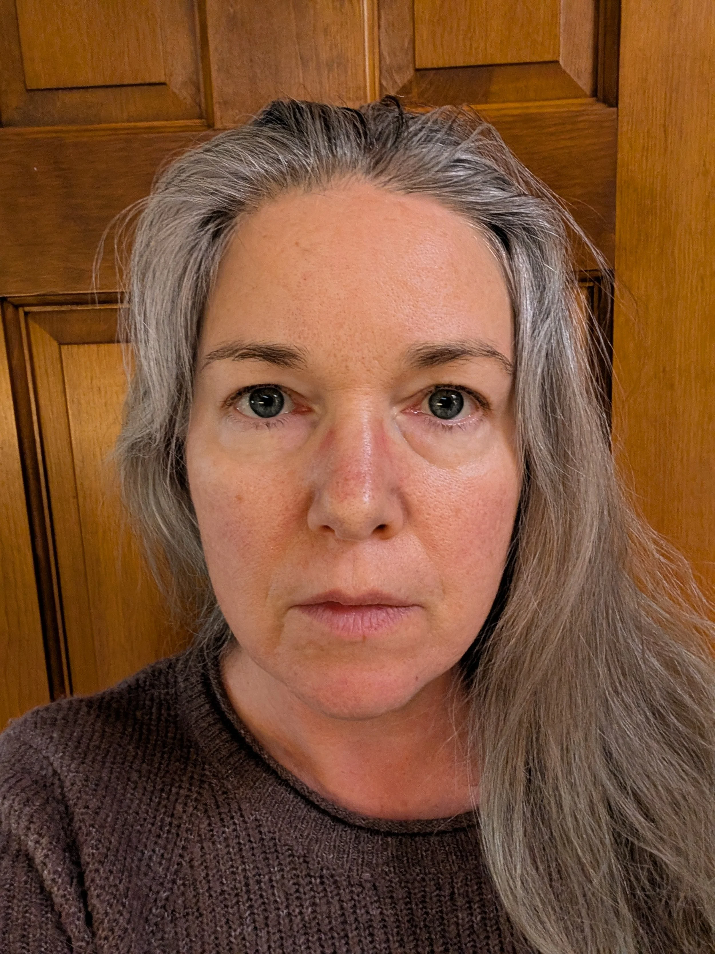 A close-up photo of a woman with gray hair, blue eyes, and freckles, wearing a brown sweater, standing in front of a wooden door.