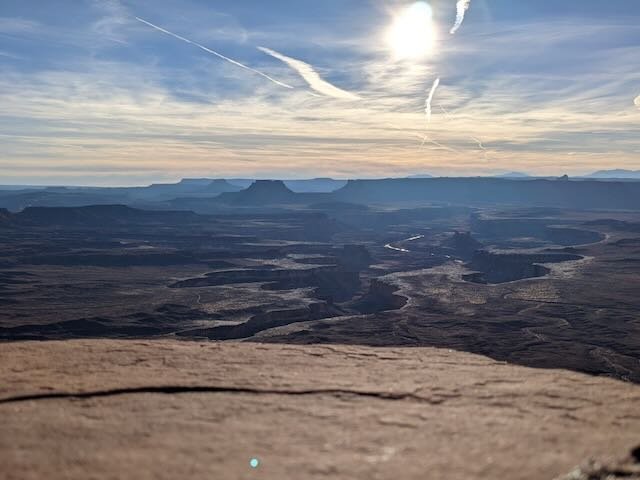A vast desert landscape with mesas and canyons under a blue sky with streaky clouds and the sun.