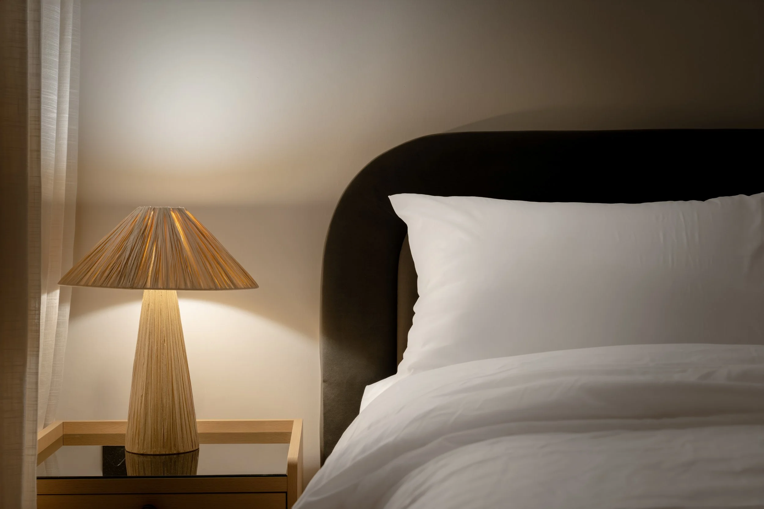 A bedroom corner with a bed, white pillow, beige wall, and a wooden side table with a tall conical lampshade.