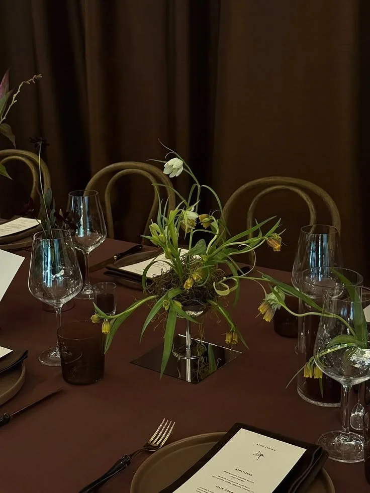 Elegant dining table set with wine glasses, cutlery, black napkins, a floral centerpiece, and a menu card, with a dark background and brown tablecloth.