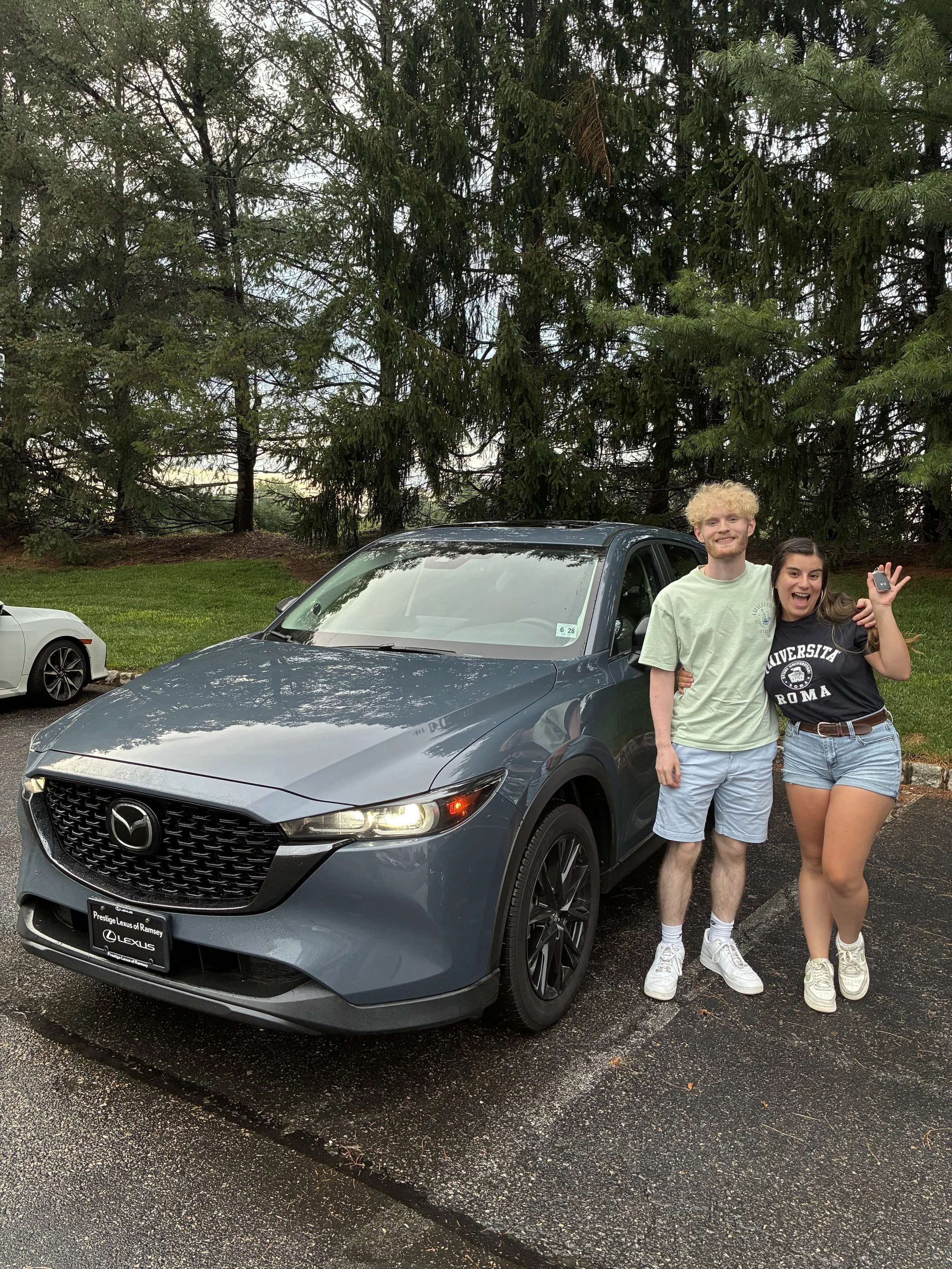 A young man and woman are standing together next to a gray Mazda SUV in a parking lot with wet pavement. They are smiling and appear happy. The woman is holding a small object in her hand. In the background, there is a large evergreen tree and another white car is partially visible on the left.