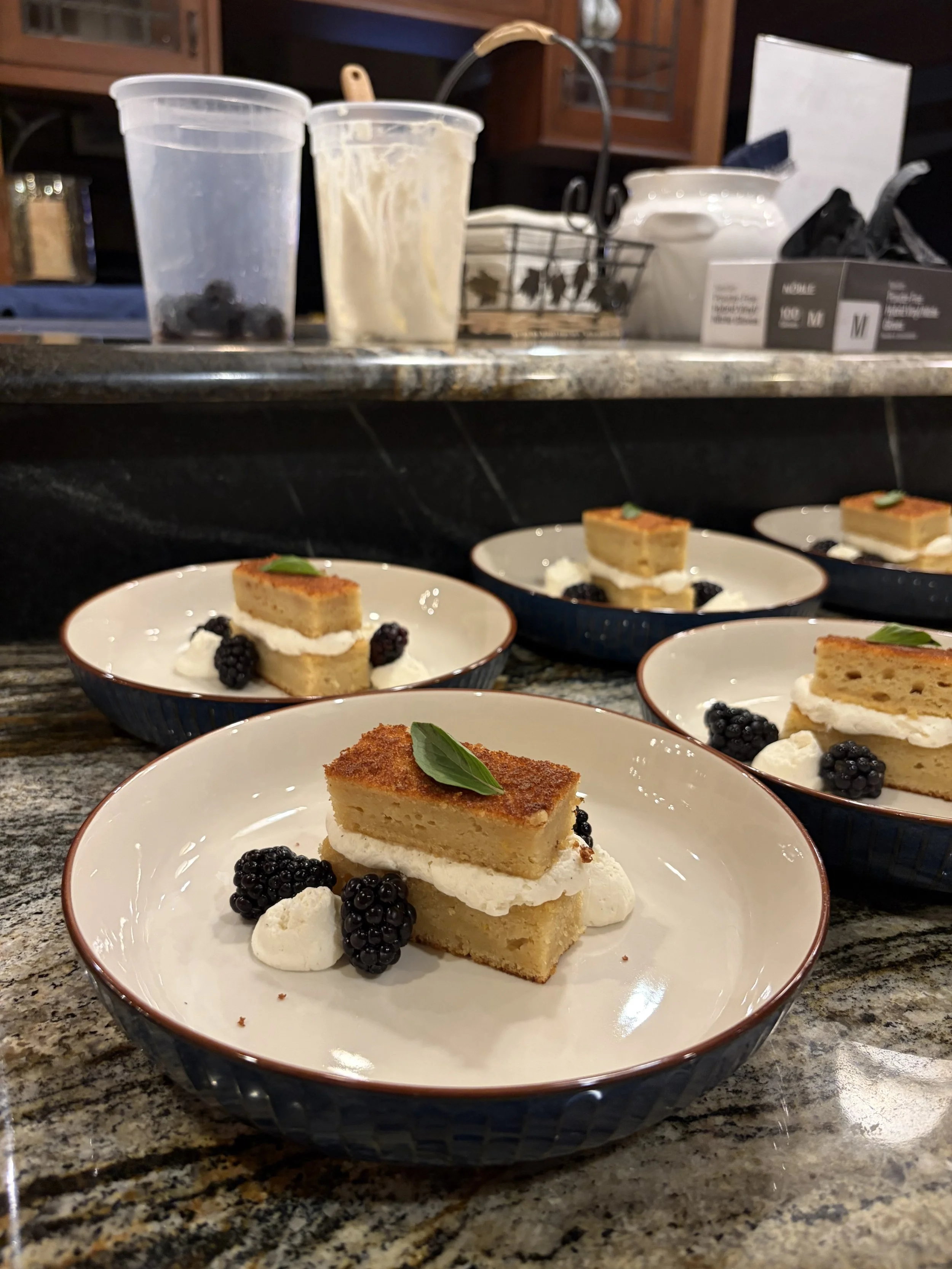 Several plates of dessert featuring slices of cake with whipped cream and blackberries on a granite countertop in a kitchen setting.