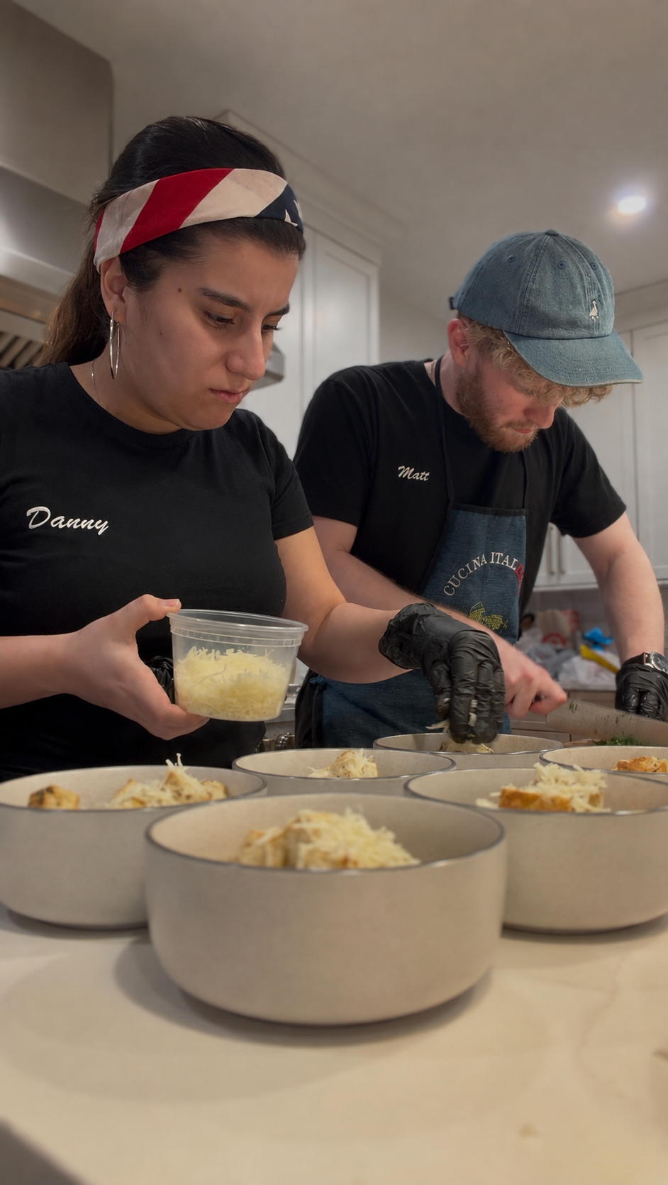 Two people, a woman with a red, white, and blue headband and a man with a denim cap, are preparing bowls of food in a kitchen. They are wearing black shirts, the woman's shirt reads 'Danny,' and the man's reads 'Matt.' They are adding cheese to the bowls, which appear to contain lasagna or a similar dish.