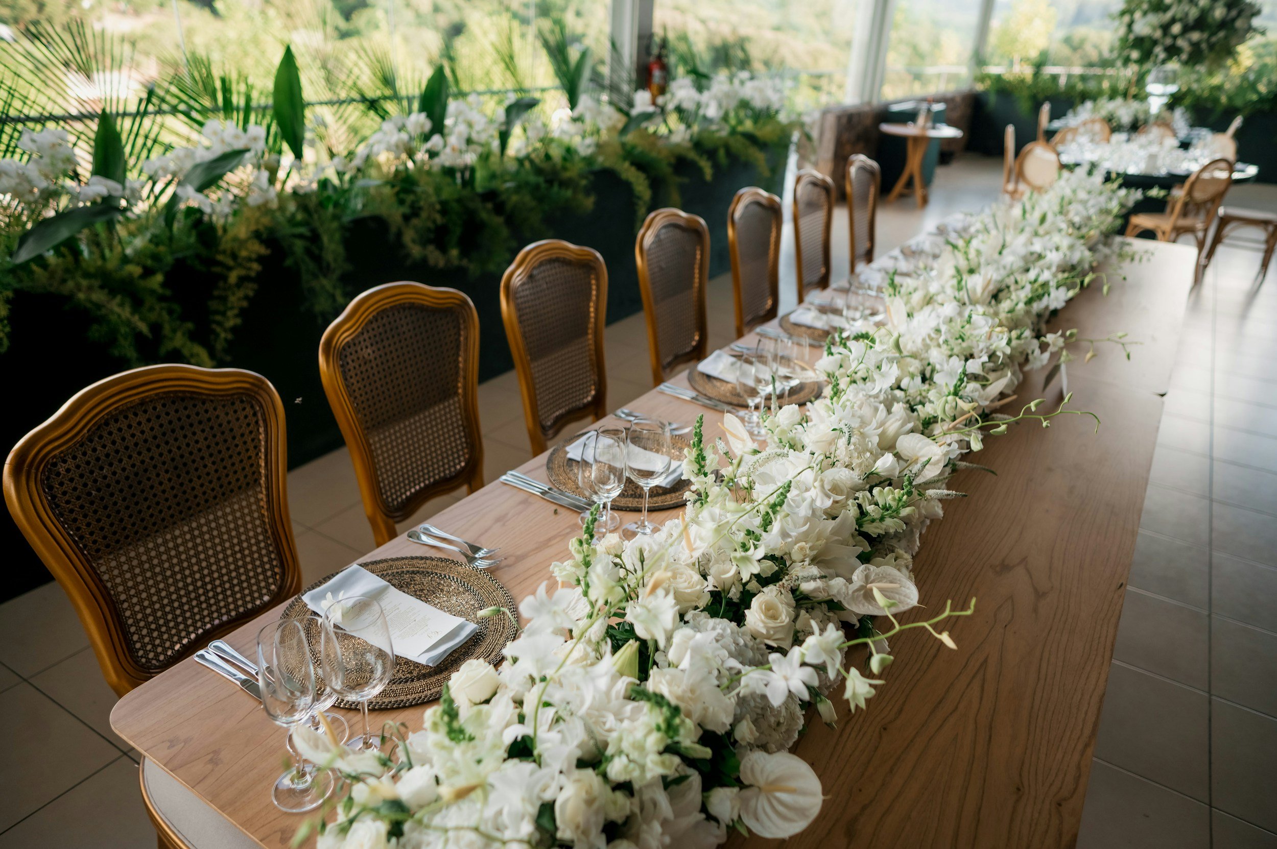 A long banquet-style table decorated with white flowers and greenery, set with glassware, napkins, and plates, in a bright room with large windows and view of trees outside.