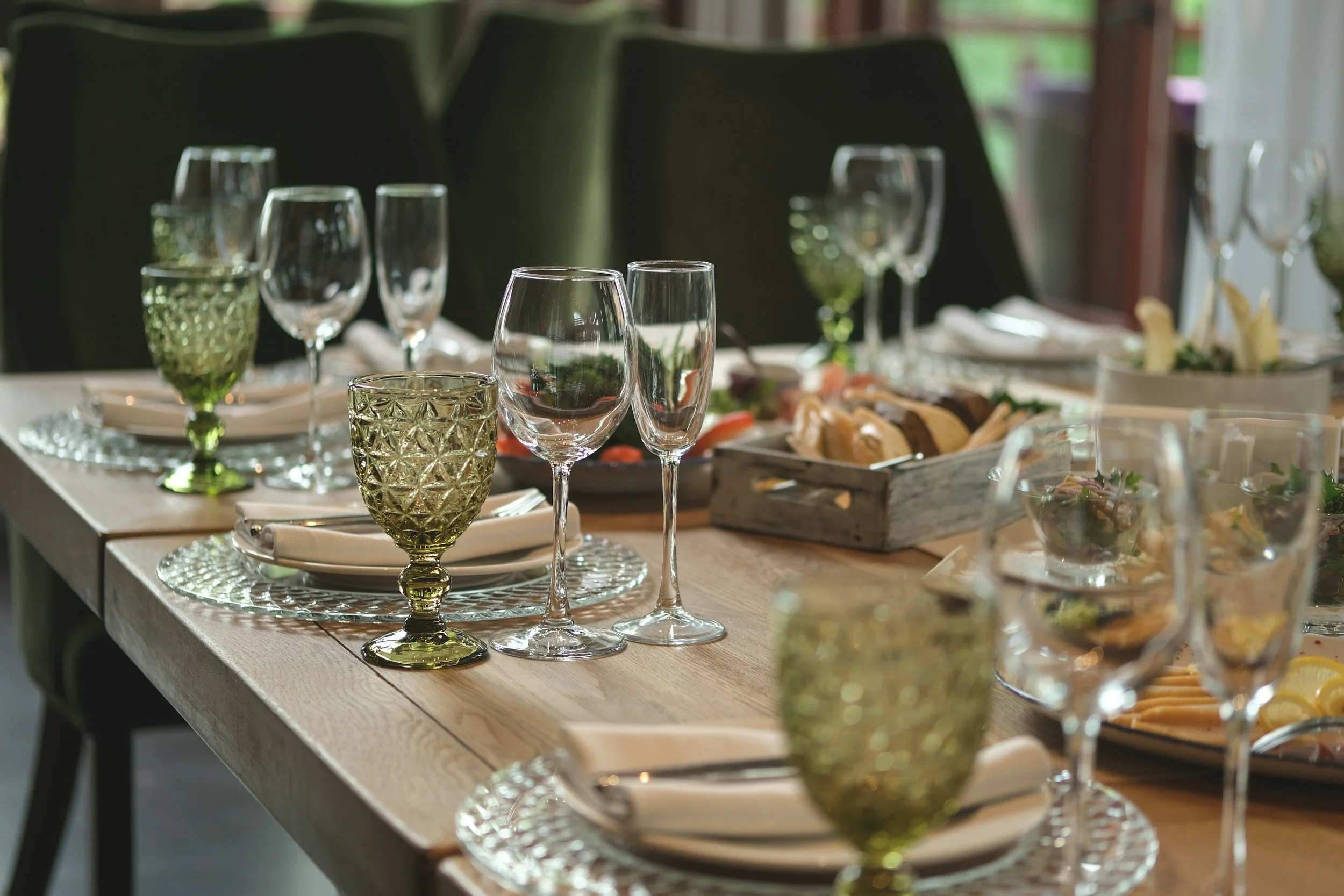 A dining table set with multiple wine glasses, glasses with green stems, and a wooden tray with assorted snacks. The table has plates with napkins and silverware, and the setting is in a well-lit room with large windows showing greenery outside.