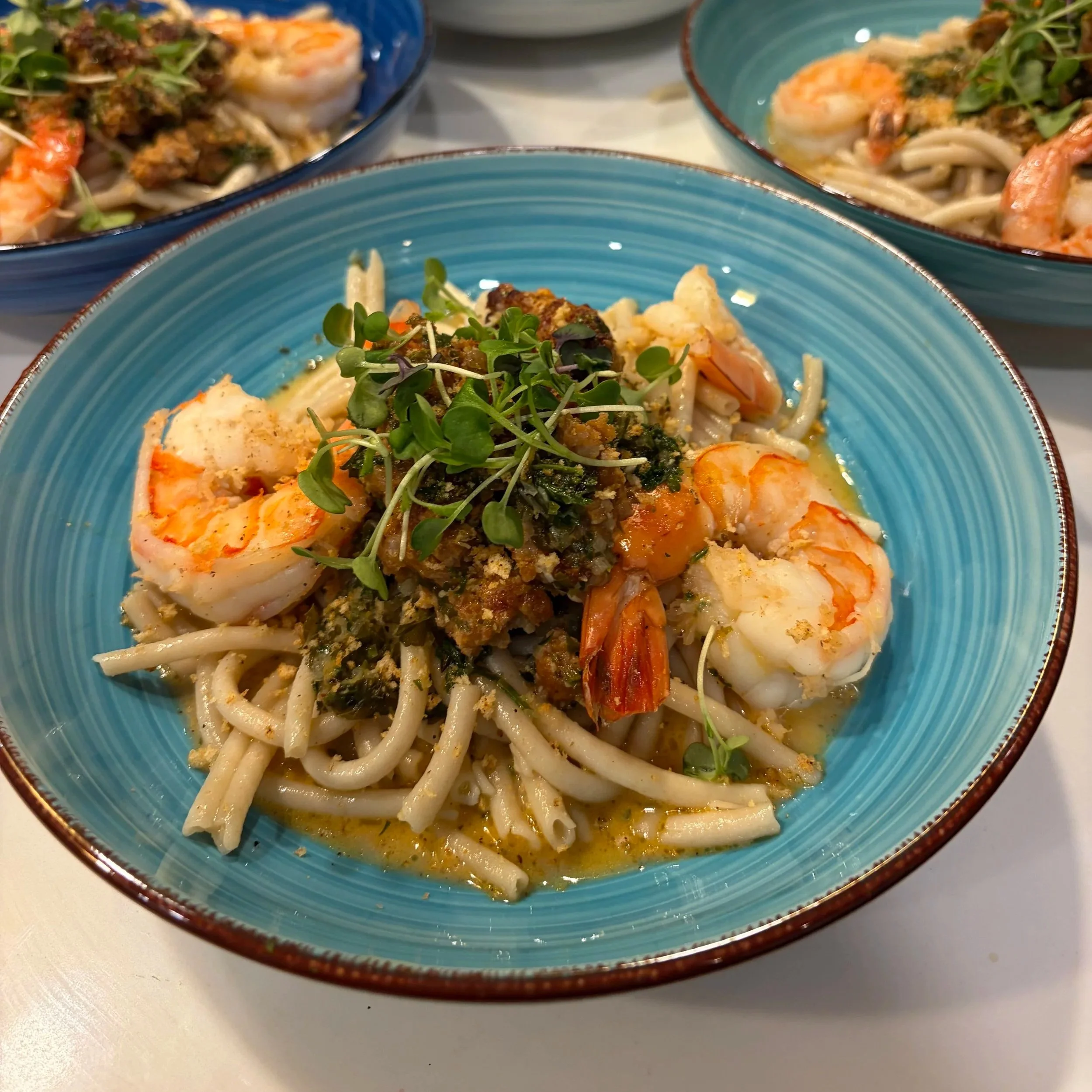 Close-up of a bowl of seafood pasta garnished with microgreens, with other bowls of similar dishes in the background.