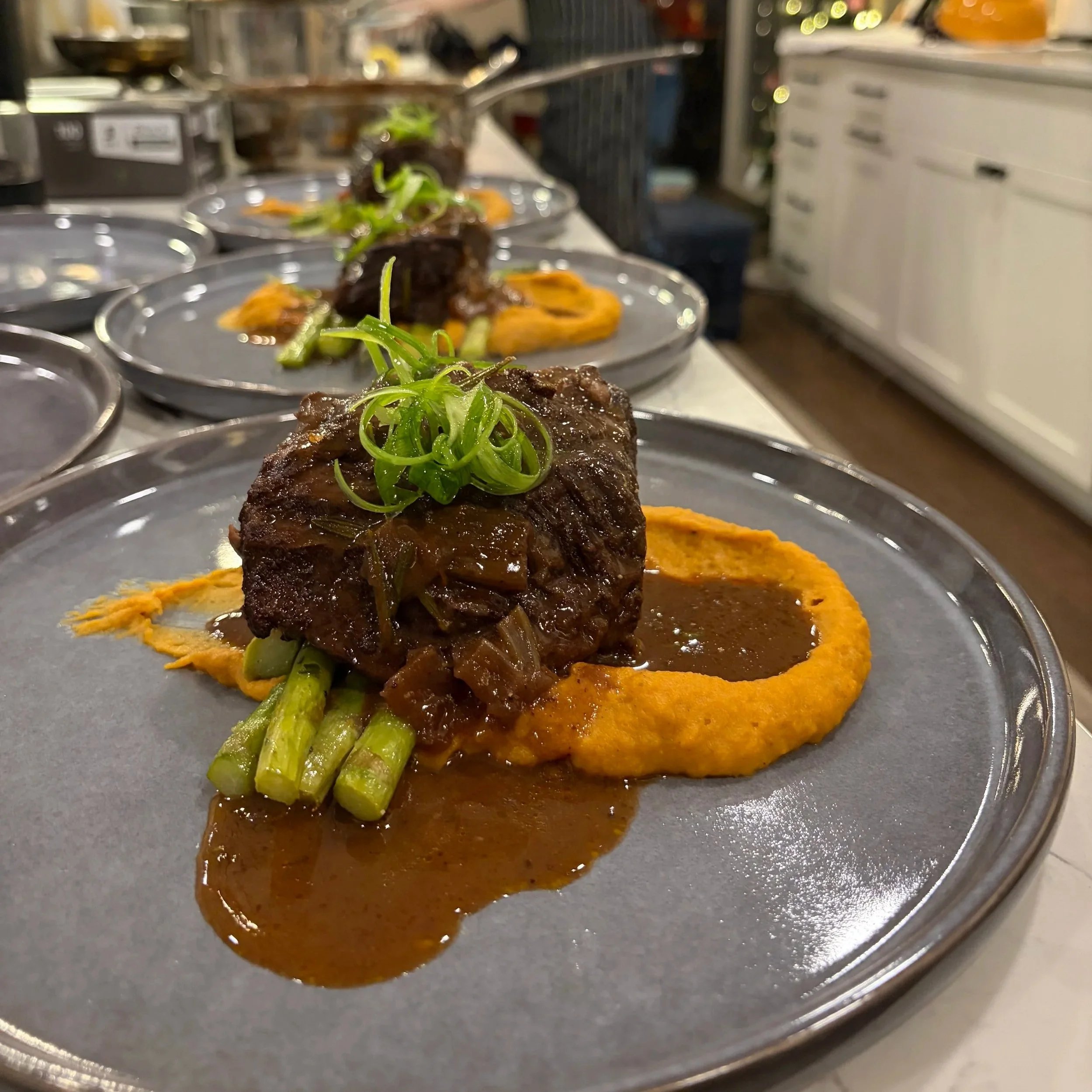 Close-up of beef short rib with sauce and green garnish on a plate, with sweet potato puree underneath, in a commercial kitchen.